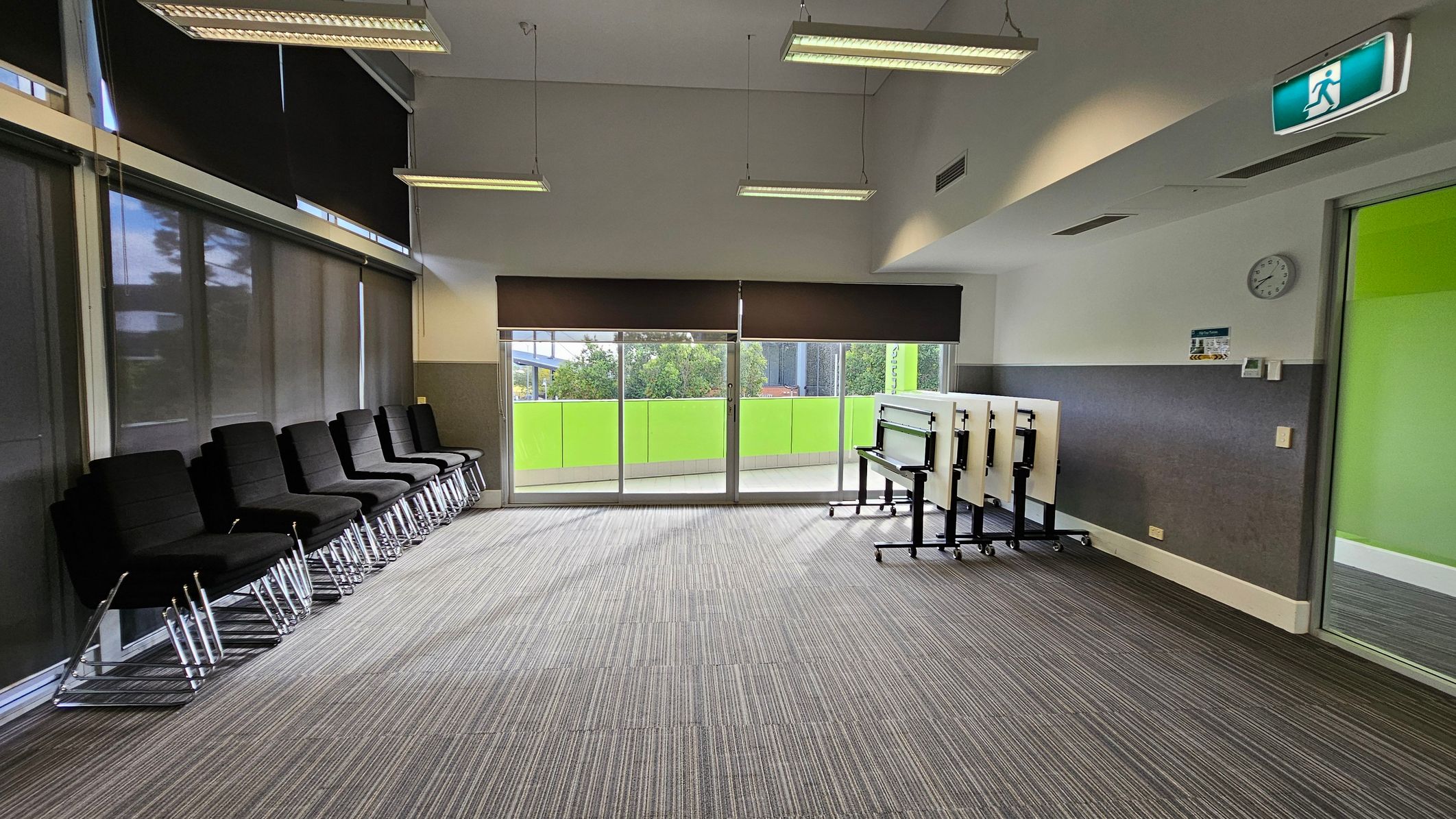 A room with a grey carpeted floor and a white ceiling featuring hanging lights. On the left, there are black chairs lined up against a wall with dark blinds. The right side has a green wall with a clock and a stack of folded tables on wheels. The back wall has large windows with a view of greenery outside, partially covered by blinds. An exit sign is visible near the ceiling.