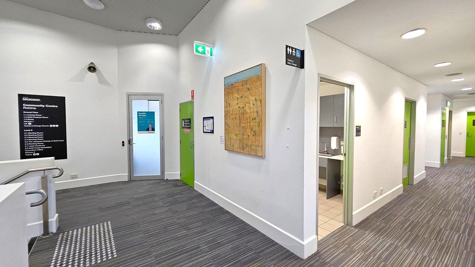 A hallway with grey carpet and white walls. On the left, a black sign lists the facilities of the Gold Coast Community Centre Robina. A security camera is mounted above. A green door and a glass door with a poster are visible. In the middle, a large abstract painting hangs on the wall. On the right, there are open doorways leading to a bathroom and other rooms, with green doors further down the corridor. The ceiling has recessed lighting and an emergency exit sign.