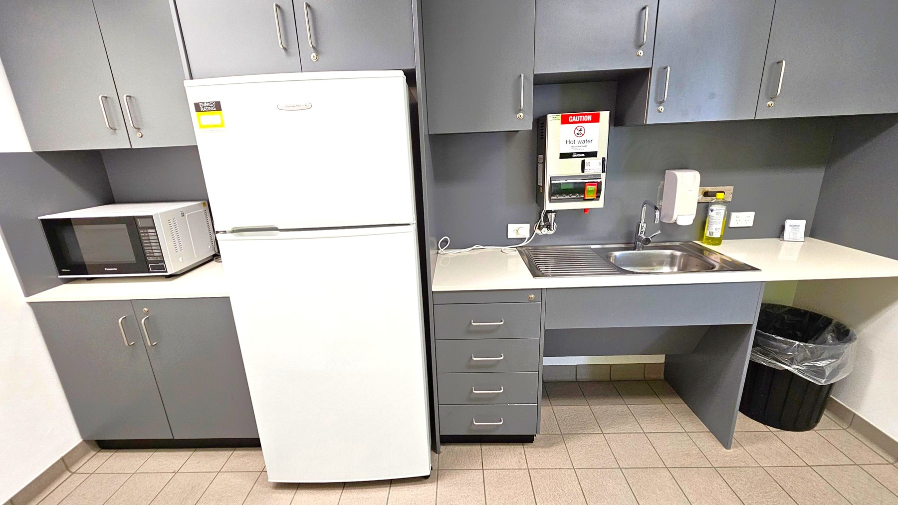 A kitchen area with grey cabinets and a tiled floor. On the left, a microwave sits on a countertop beneath wall cabinets. Next to it is a white refrigerator. In the centre, a sink with a draining board is set into the counter, with a hot water dispenser above. To the right, a soap dispenser and cleaning supplies are on the counter. Below, there are drawers and a black rubbish bin with a liner. The ceiling is not visible.