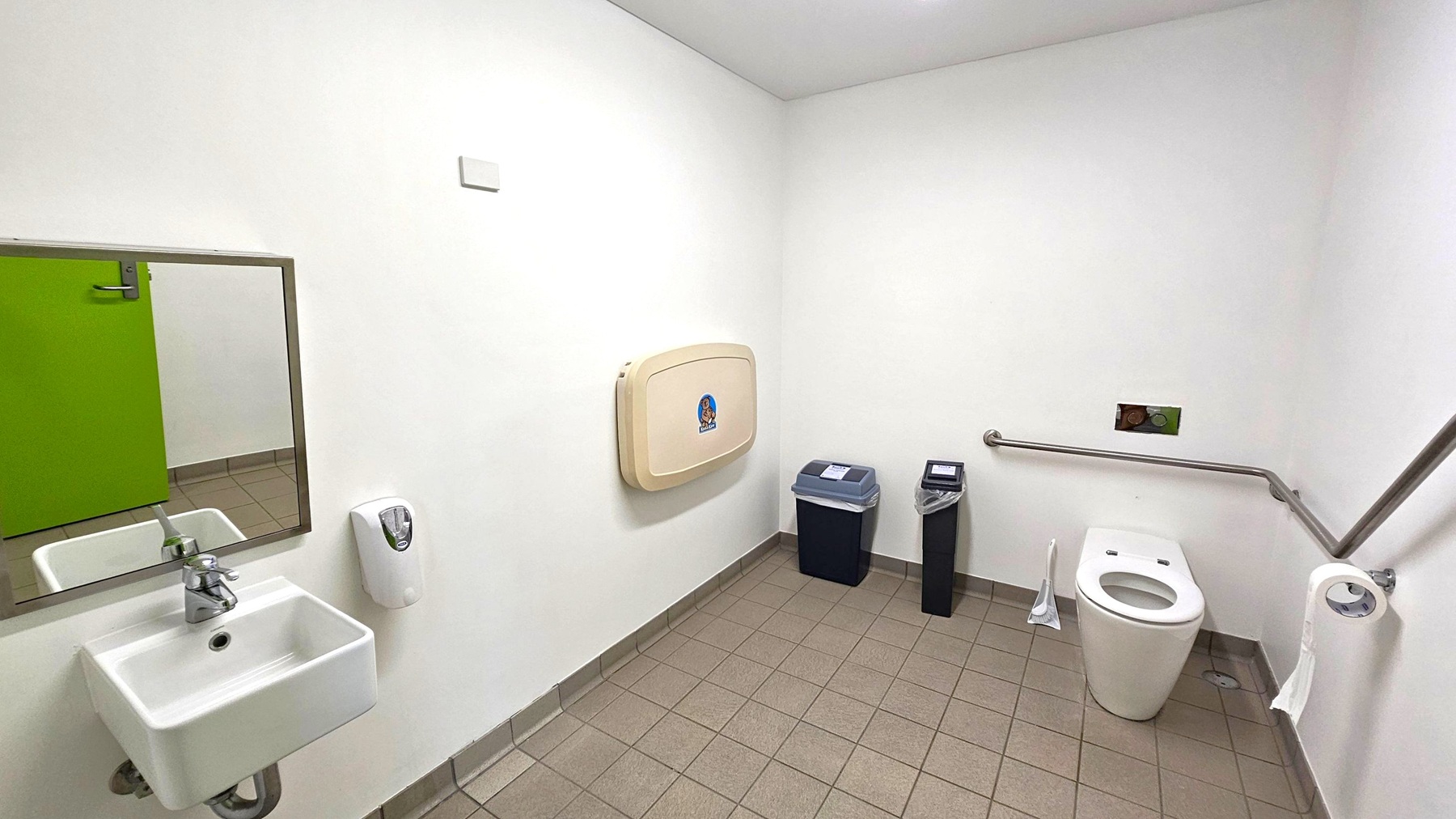 A public restroom with a white sink and mirror on the left. Below the mirror is a soap dispenser. The floor is tiled in a light brown colour. On the right, there is a white toilet with a grab bar beside it and a toilet roll holder. A baby changing station is mounted on the wall above two bins, one with a blue lid and the other with a black lid. The walls and ceiling are white, and a green door is partially visible in the mirror.