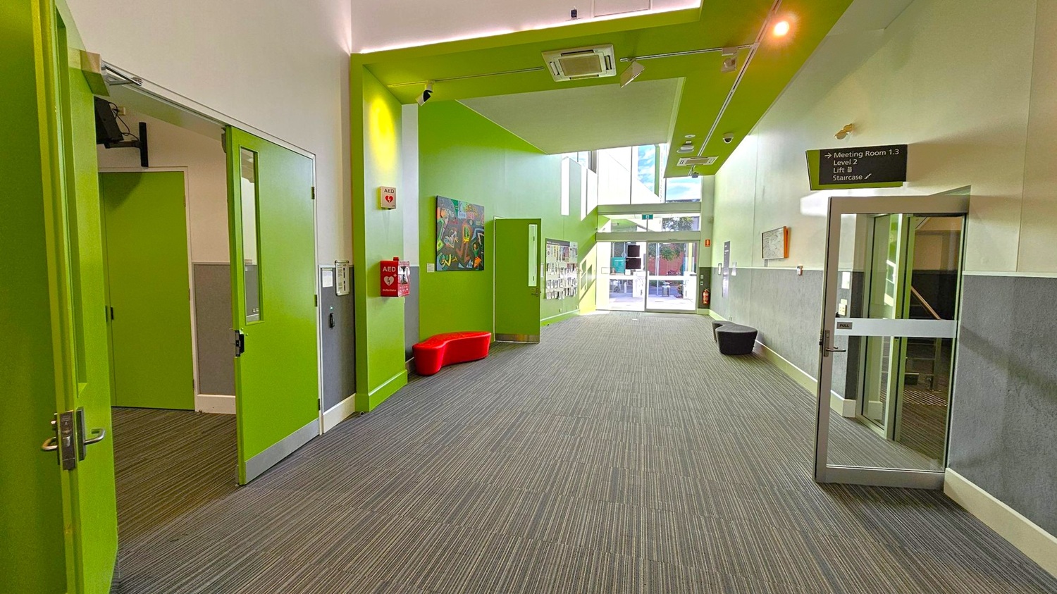 A hallway with bright green walls and doors. On the left, there are two green doors, one slightly open. The floor is covered with grey carpet tiles. In the centre, a red bench is placed against the wall, with a fire extinguisher and an AED mounted above it. The right side has a glass door with a sign above indicating directions to meeting rooms and stairs. The ceiling features green panels and recessed lighting. At the far end, large windows allow natural light to enter.