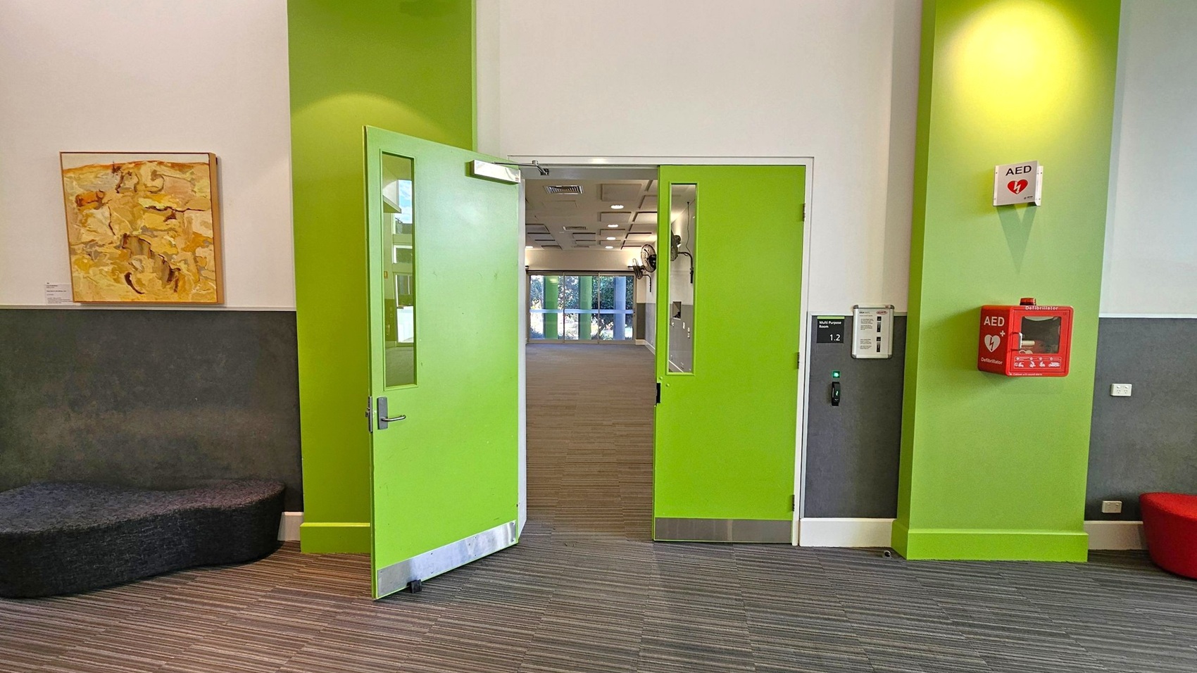 Bright green double doors are in the centre, slightly open, leading to a room with large windows. On the left, a grey bench sits below a framed abstract painting on a white and grey wall. On the right, a green wall features a red AED box and a small sign above it. The floor is covered with striped carpet tiles, and the ceiling has recessed lighting.