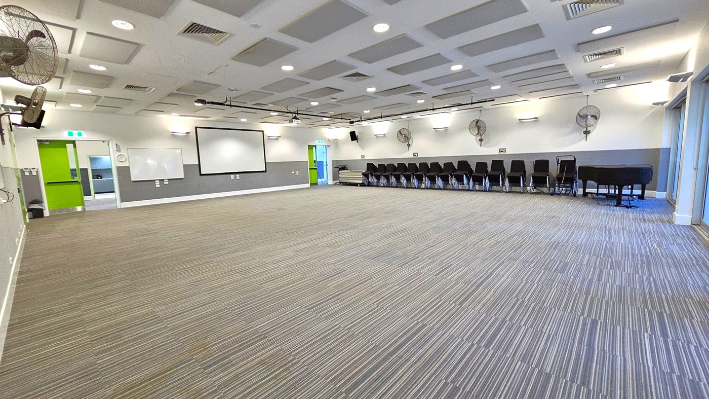 A spacious room with a striped carpeted floor. On the left, there are green double doors and a whiteboard mounted on the wall. In the centre, a large projection screen is visible. On the right, a row of stacked black chairs lines the wall, next to a black piano. The ceiling features recessed lighting and ventilation panels, with wall-mounted fans along the right side. The room is well-lit with a modern design.