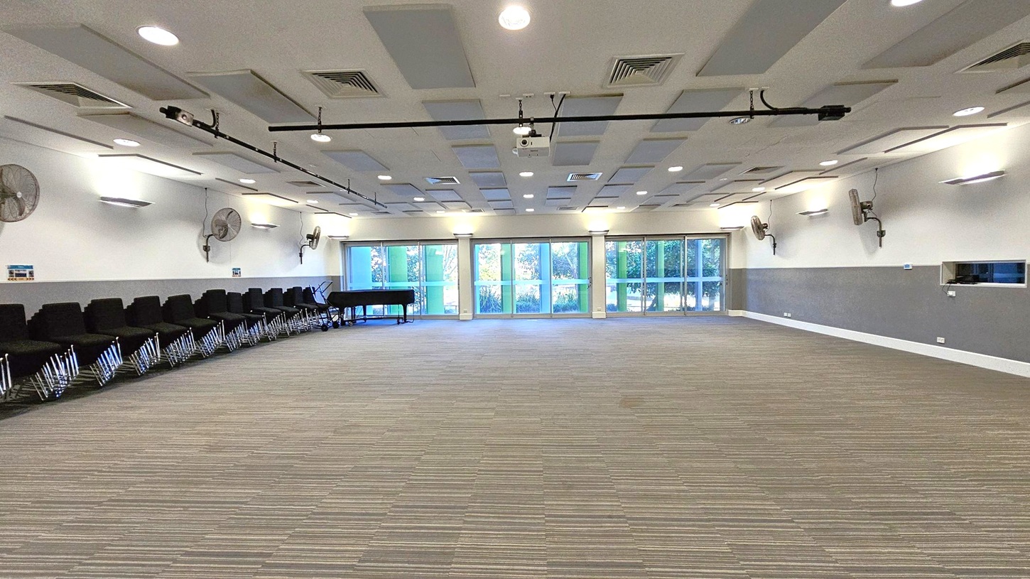 A spacious room with a striped carpeted floor. On the left, there are rows of black chairs lined up against the wall, with a black grand piano nearby. The right wall features several wall-mounted fans and lights. The back wall is mostly glass, with large windows and doors offering a view of greenery outside. The ceiling is fitted with recessed lights and air vents, and there is a projector mounted in the centre.