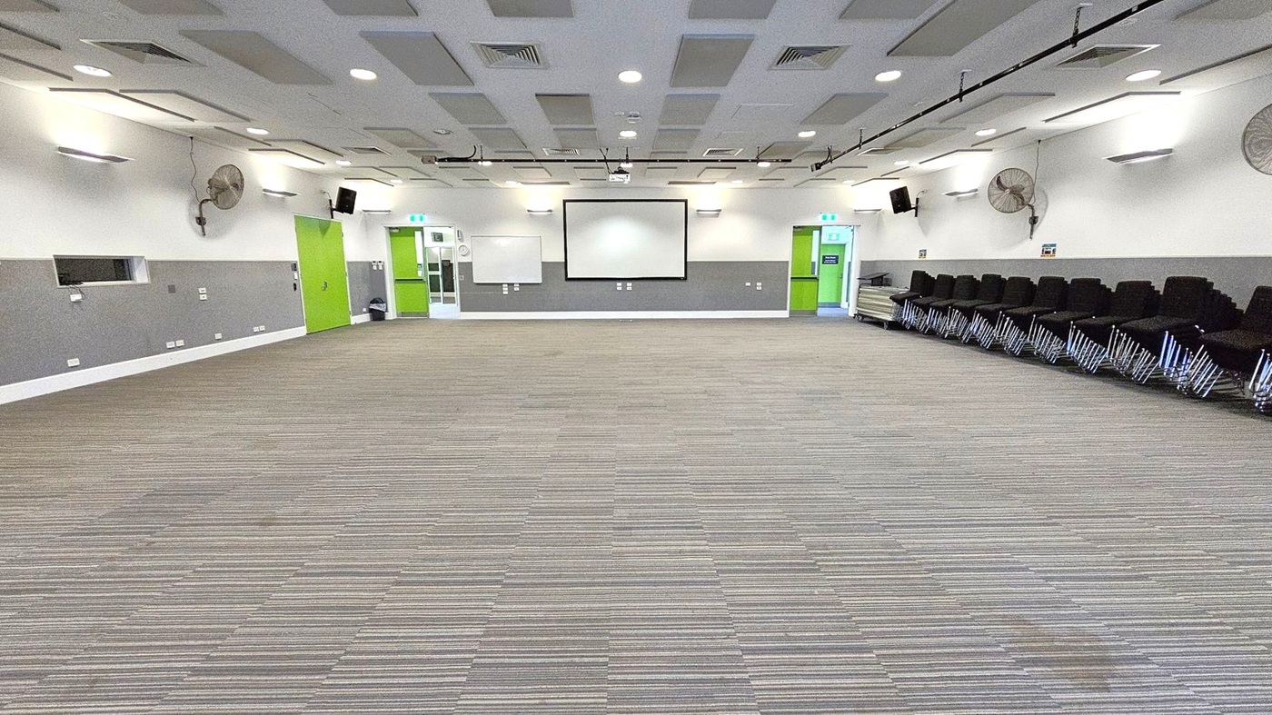 A spacious room with a striped carpeted floor. The back wall features a large white projection screen flanked by two green doors. On the right, there is a stack of black chairs with chrome legs. The ceiling is fitted with recessed lighting and acoustic panels. The walls are grey and white, with mounted fans and speakers.