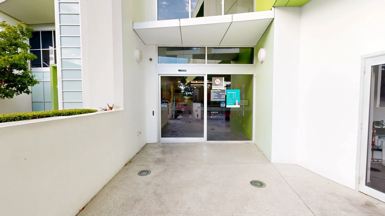A modern entrance with glass sliding doors in the centre, featuring a sign and a no-smoking symbol. The building exterior is white with green accents. On the left, there's a small tree and a row of windows. The floor is concrete with two circular drains. The ceiling is white with recessed lighting. On the right, part of another glass door is visible.