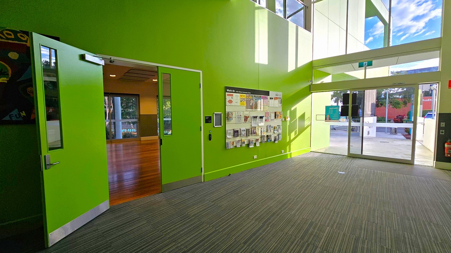 A bright interior space with lime green walls. On the left, a green door is open, revealing a room with wooden flooring. In the centre, a wall-mounted display holds various brochures and pamphlets. To the right, large glass doors lead outside, allowing natural light to flood in. The floor is covered with grey carpet tiles, and the ceiling is not visible.