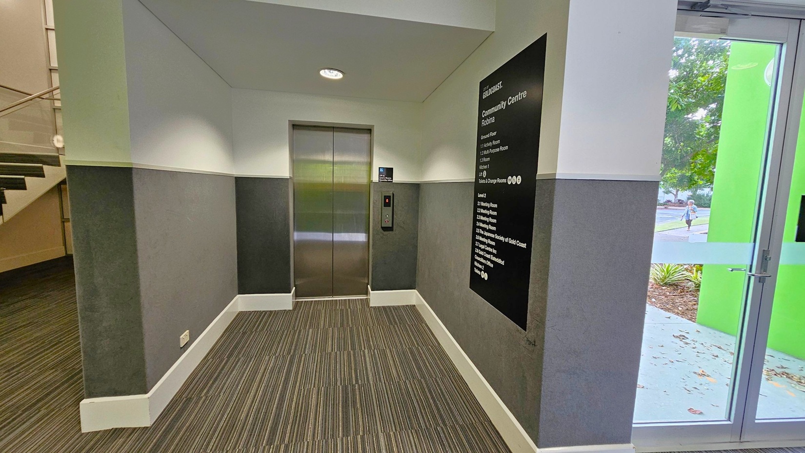 A hallway with a lift in the centre, featuring metallic doors. To the right, a black directory sign lists various rooms and facilities. A glass door with a green frame is also on the right, showing a view of the outside with trees and a person walking. On the left, a staircase with a wooden handrail is partially visible. The floor is covered with striped carpet, and the walls are painted in two tones of grey. The ceiling is plain with a recessed light.