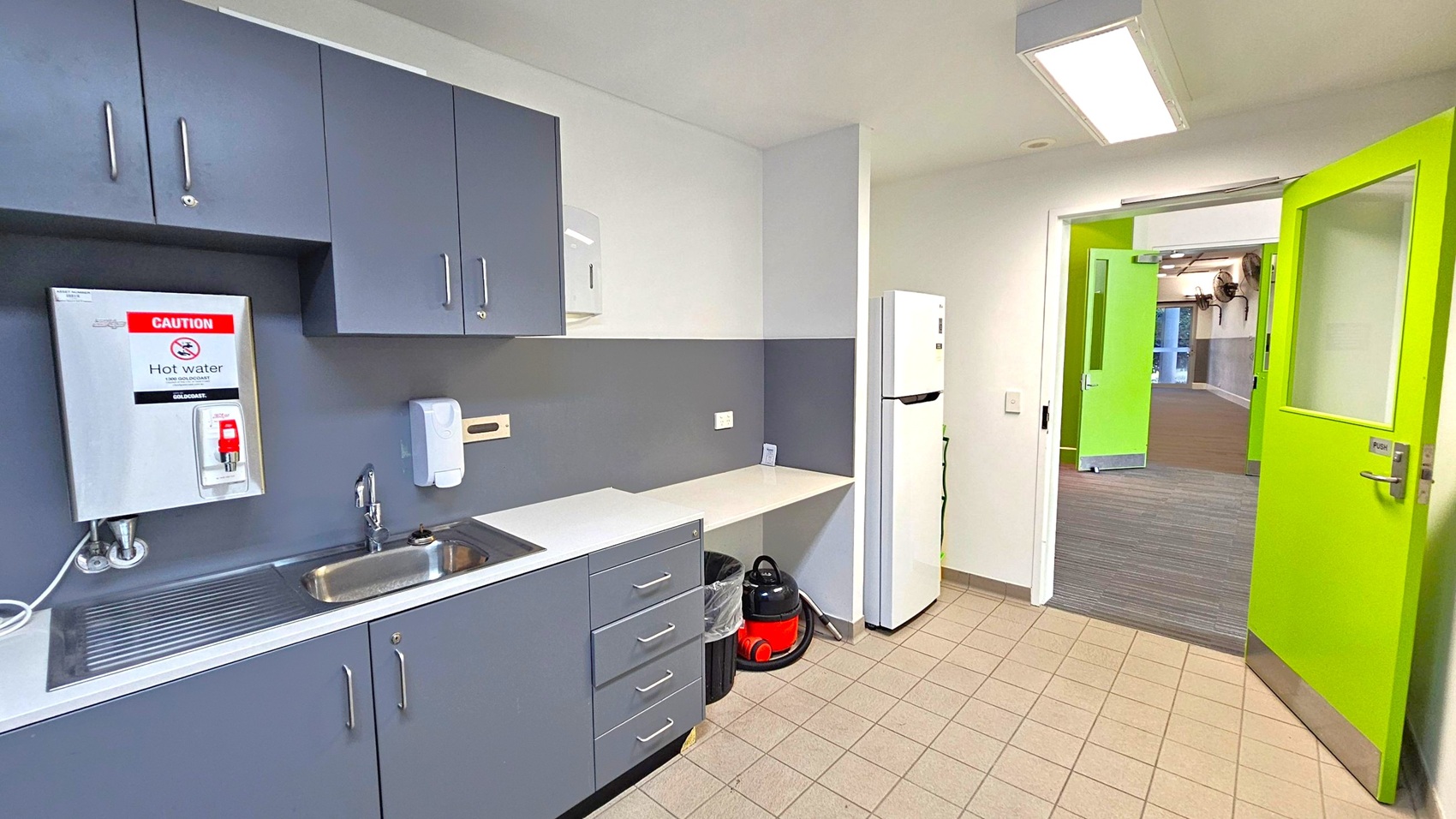 A small kitchen area with grey cabinets and a stainless steel sink on the left. Above the sink is a hot water dispenser with a caution sign. Below the counter are drawers and a bin. A white refrigerator stands to the right, next to a small counter space. A red and black vacuum cleaner is on the floor. The floor is tiled, and the ceiling has a fluorescent light. A bright green door is open, leading to a carpeted hallway with another green door visible.