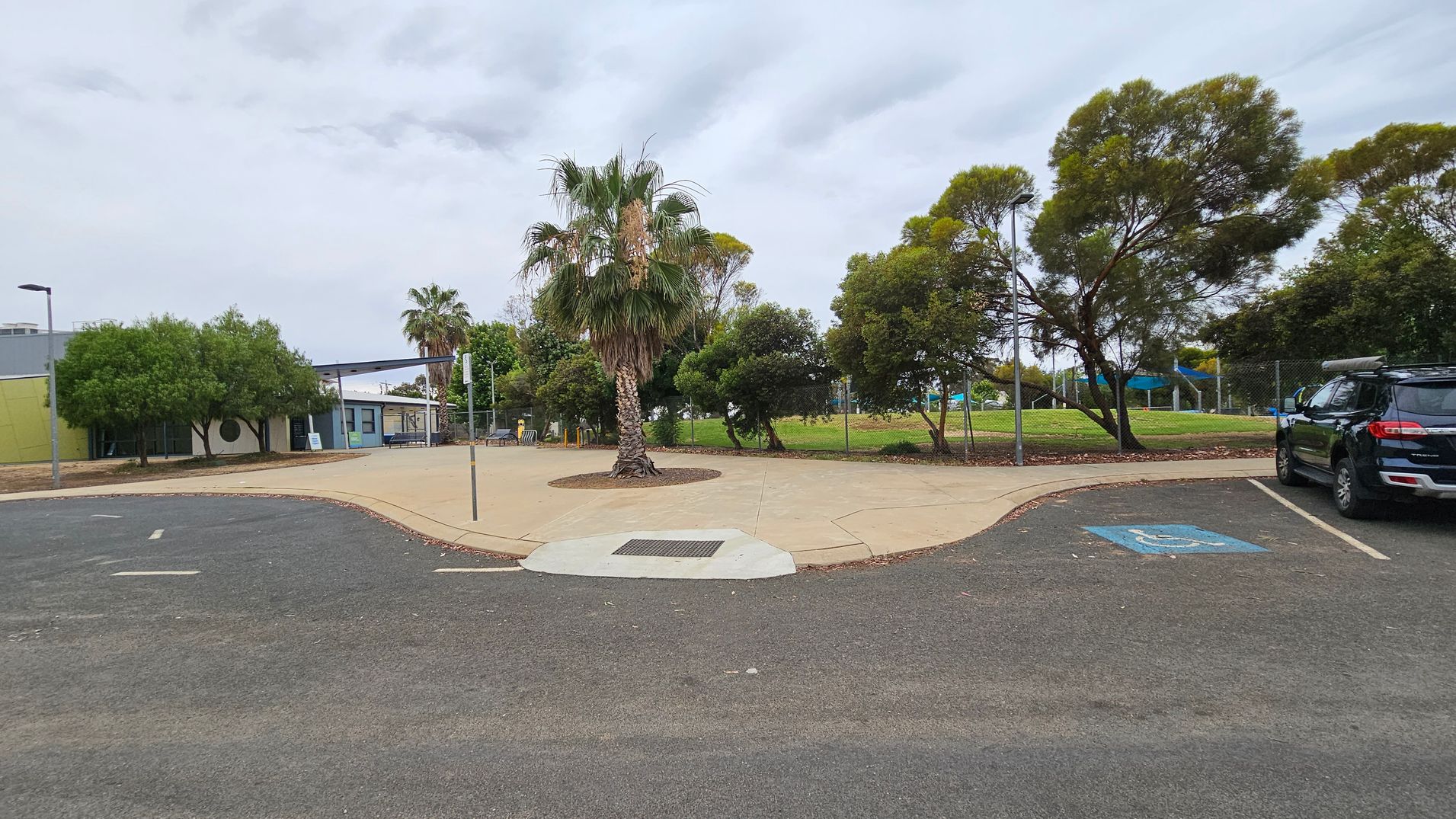 A car park with a marked disabled parking space on the right. A black SUV is parked nearby. In the centre, a palm tree is surrounded by a paved area. To the left, a building with a light green wall and a few windows is partially visible, with trees in front. In the background, a grassy area with more trees and a playground with blue shade structures is enclosed by a chain-link fence. The ground is a mix of asphalt and concrete paving.