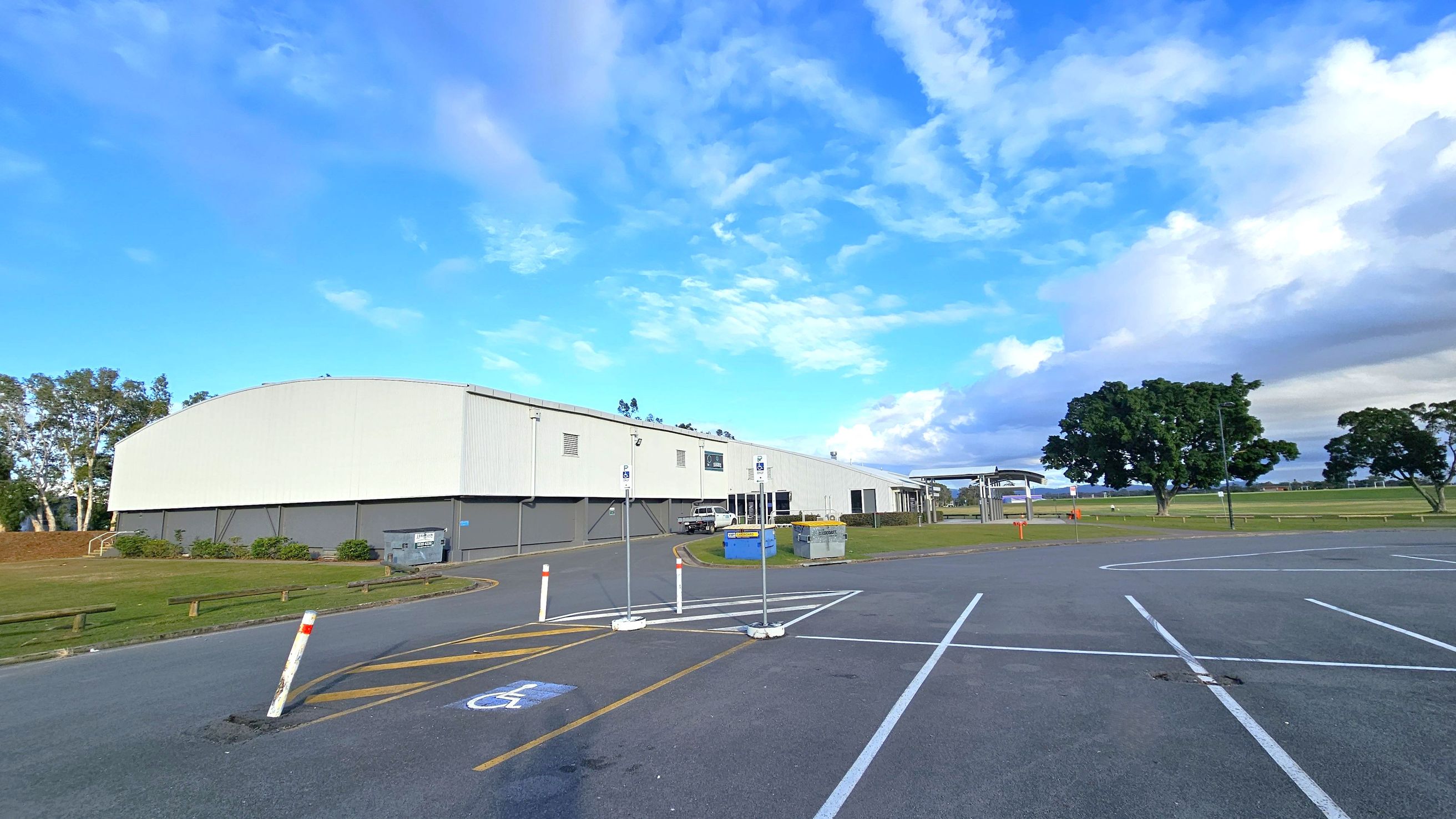 A large, white industrial building is on the left, with a grey lower section and a few small windows. In front of it, a car is parked near a blue dumpster. The foreground features a car park with marked spaces, including a disabled parking spot. On the right, there are open grassy fields with a few large trees. The background shows more trees and a clear sky.