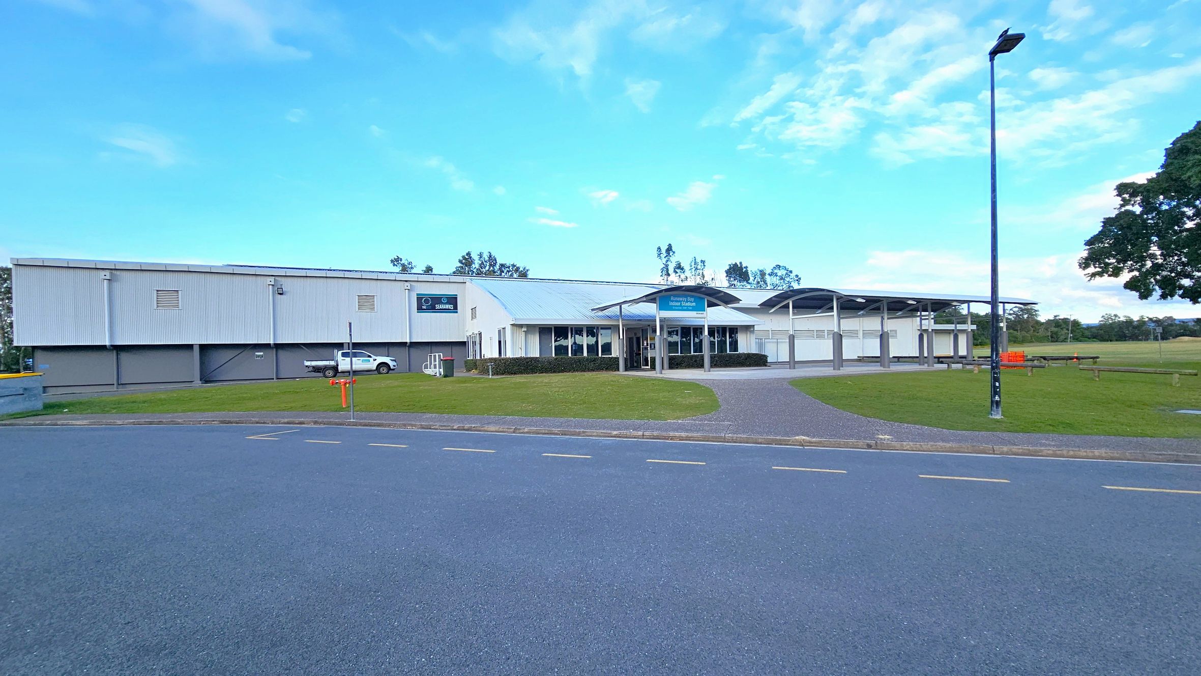 A large, modern building with a white exterior and a curved roof is situated in the centre. The entrance features a blue sign and is flanked by glass windows. A white vehicle is parked on the left side near the building. A wide, paved road runs across the foreground, with a grassy area and a pathway leading to the entrance. A tall streetlamp stands on the right, and there are trees in the background.