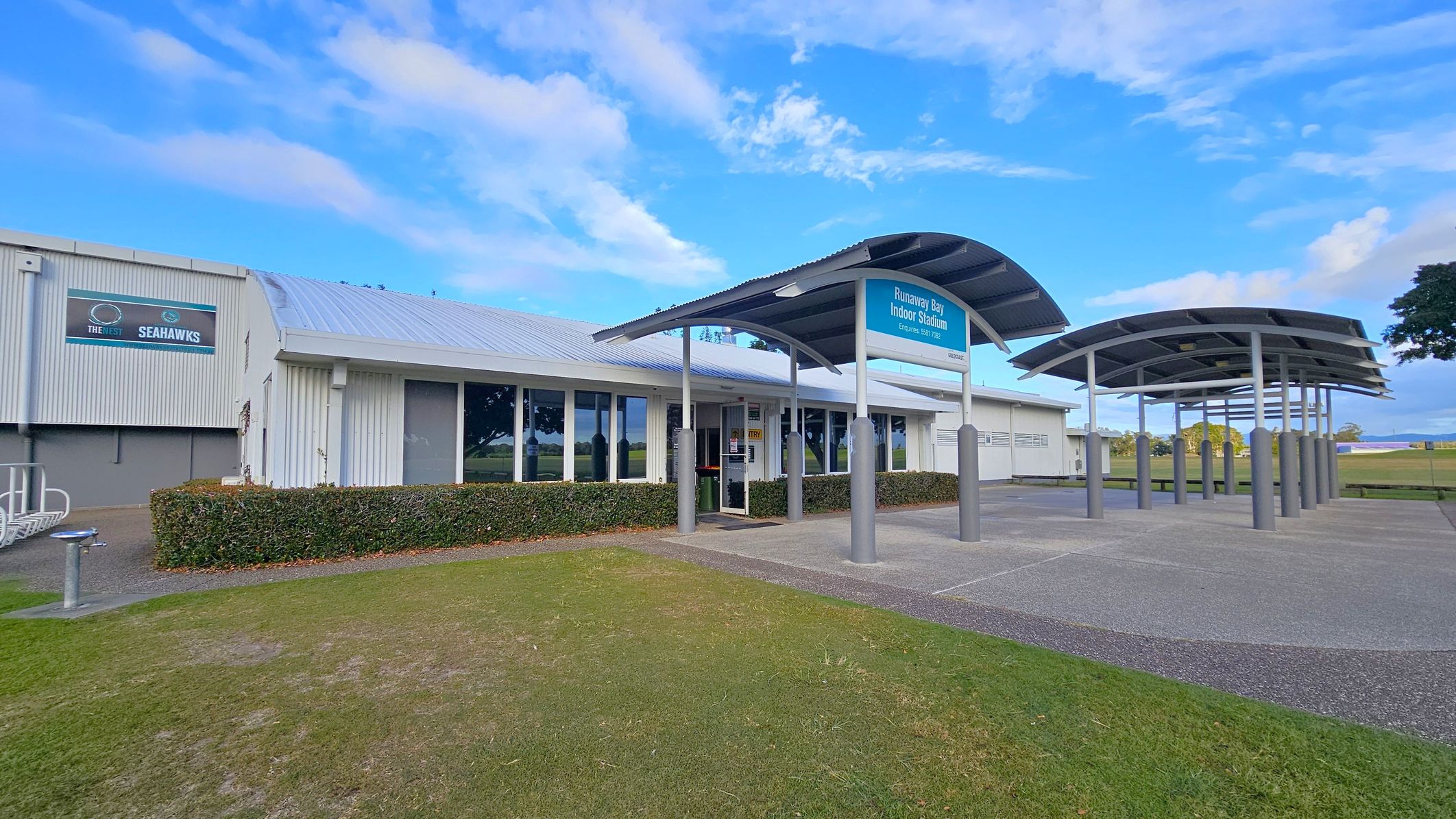 A building with a metal roof and large windows on the left, featuring a sign for "The Nest Seahawks." In the centre, a covered entrance with a sign for "Runaway Bay Indoor Stadium" supported by metal poles. The foreground has a grassy area and a paved path leading to the entrance. On the right, there are more covered structures and open space with trees in the distance.