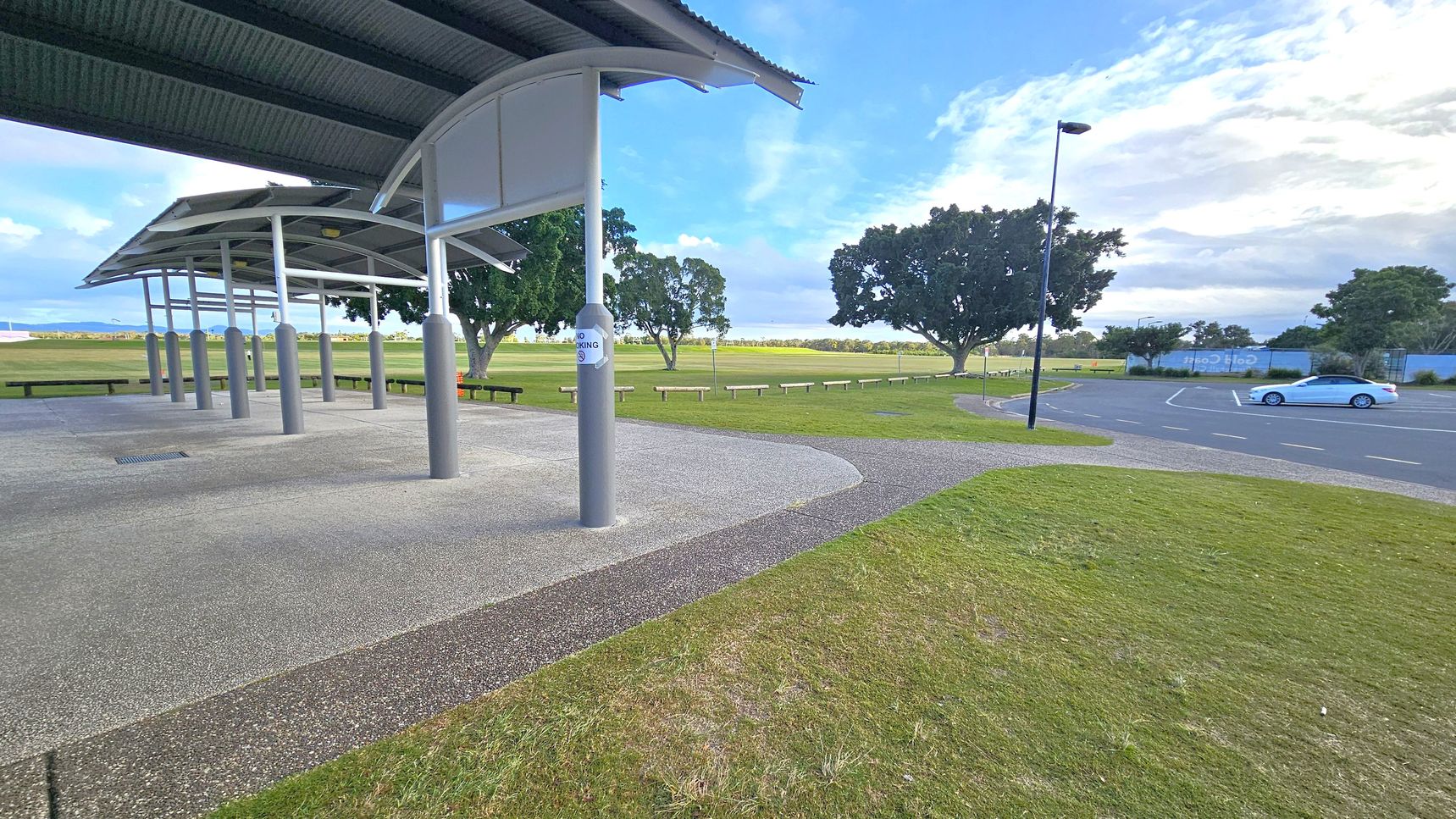 A covered area with a corrugated metal roof supported by grey pillars is on the left. The ground is paved with a textured surface. In the middle, there is a grassy area with a few trees and benches. On the right, a car park with a single parked car and a tall streetlamp. The background features more trees and a distant view of open fields.
