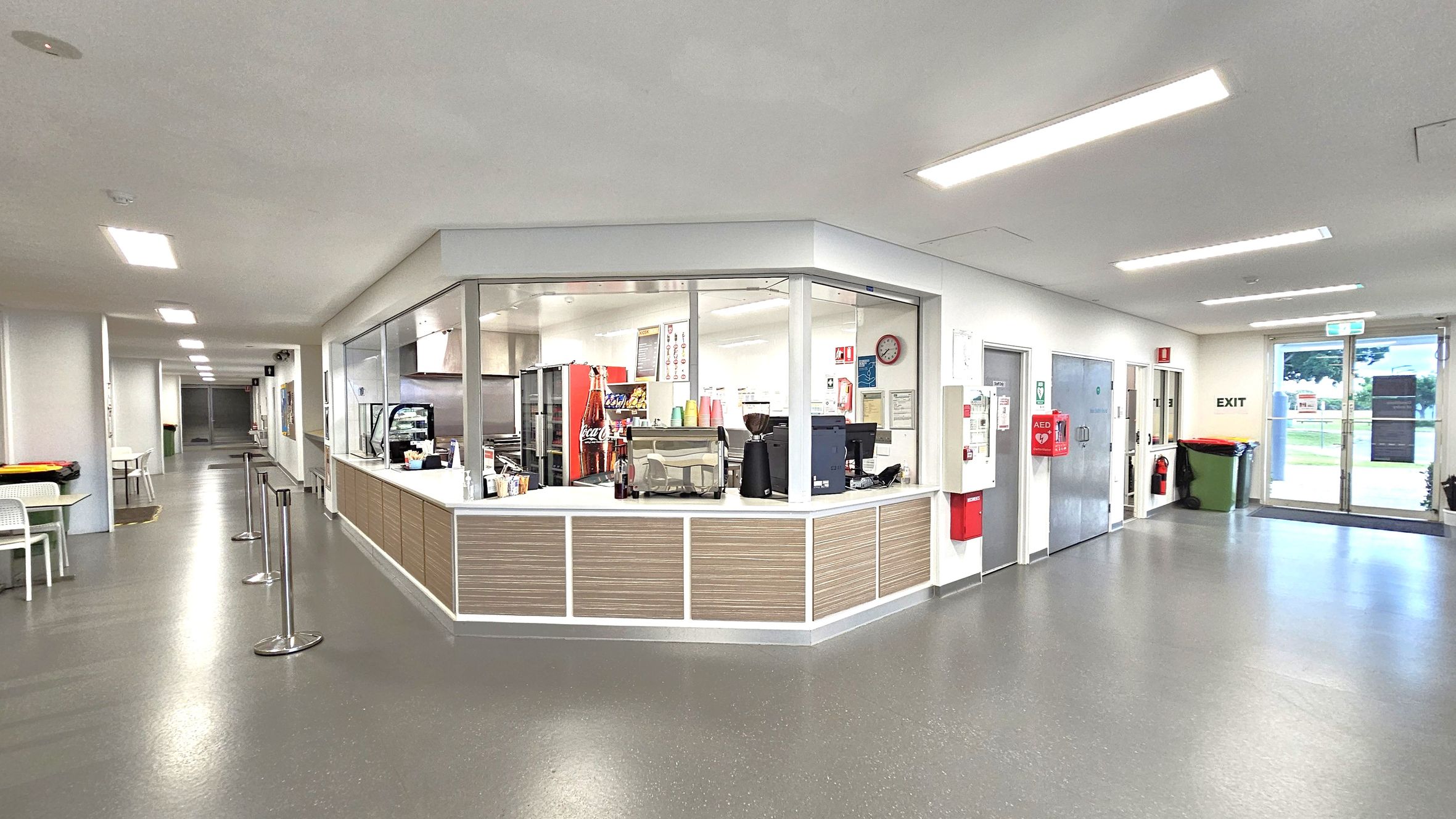A spacious indoor area with a canteen in the centre, featuring a counter with a coffee machine, snacks, and a drinks fridge. The left side has a corridor with tables and chairs along the wall. On the right, there are doors, a fire extinguisher, and a green bin near an exit. The floor is grey, and the ceiling has recessed lighting.