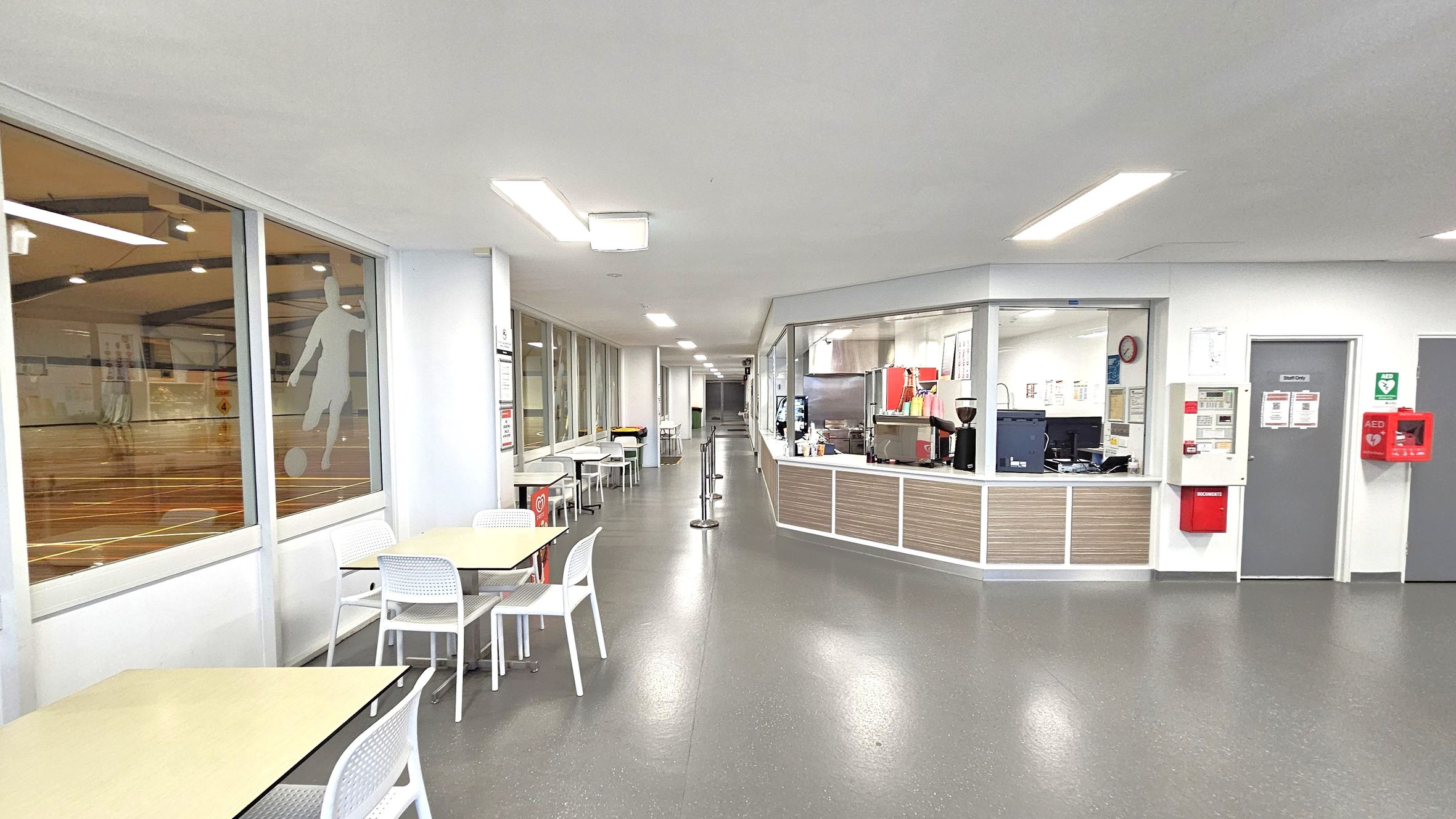 A spacious indoor area with a grey floor and white ceiling. On the left, there are large windows with a silhouette of a footballer, overlooking a sports court. Below the windows, tables and white chairs are arranged. On the right, a counter with a coffee machine and various equipment is visible, with a grey door and a red AED box on the wall. The area is well-lit with ceiling lights, and a corridor extends into the distance.