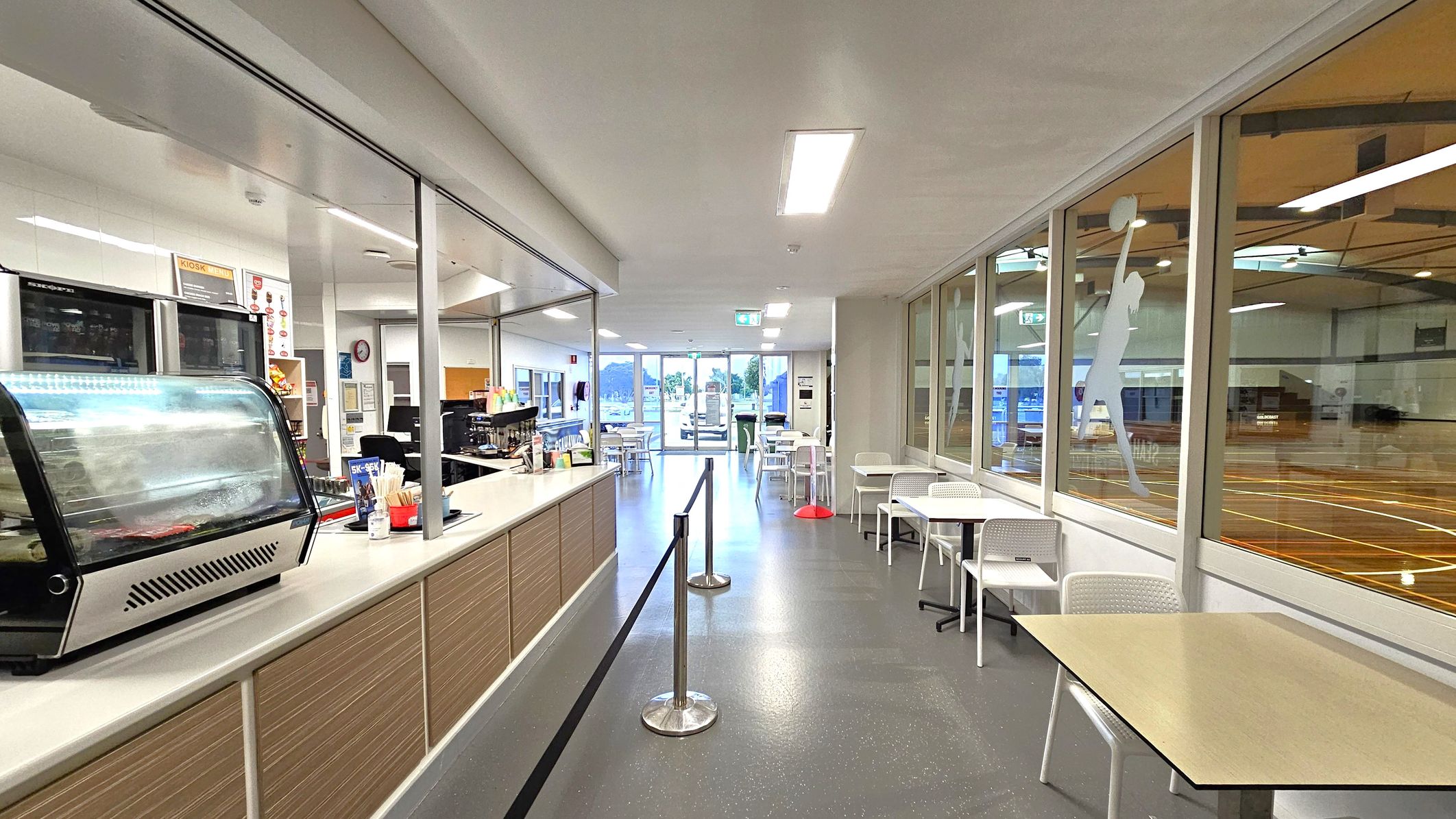 A café area with a counter on the left featuring a display case and various items. On the right, there are tables and chairs next to large windows with silhouettes of athletes. Beyond the windows, an indoor sports court is visible. The floor is grey and speckled, and the ceiling is white with recessed lighting. At the far end, glass doors lead outside, with more tables and chairs visible through them.
