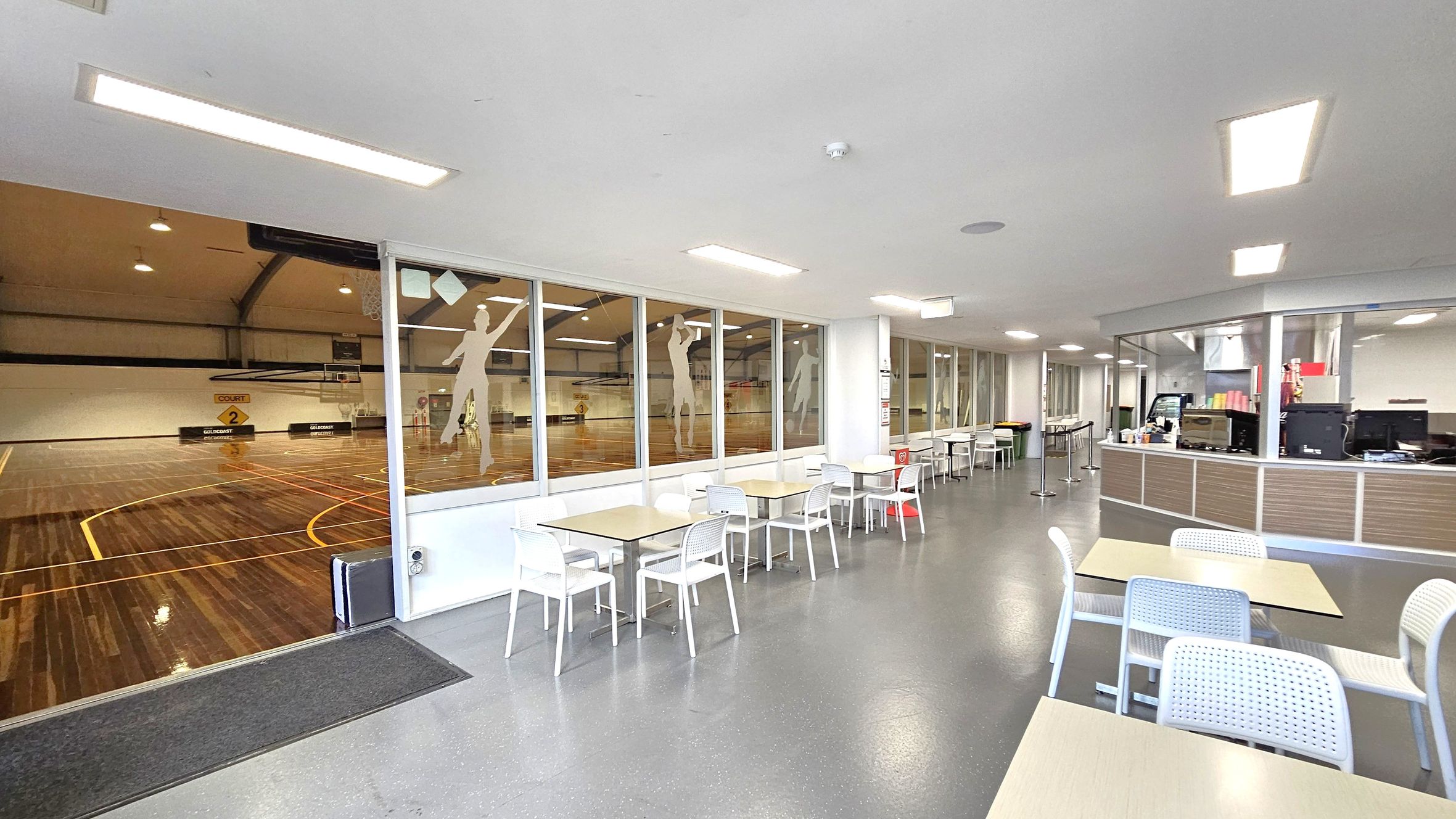 A sports facility with a basketball court on the left, visible through large glass windows. The court has a polished wooden floor with yellow lines and a sign for "Court 2". On the right, there is a seating area with tables and white chairs on a grey floor. Further right, a counter with computers and various items is visible. The ceiling is white with rectangular lights.