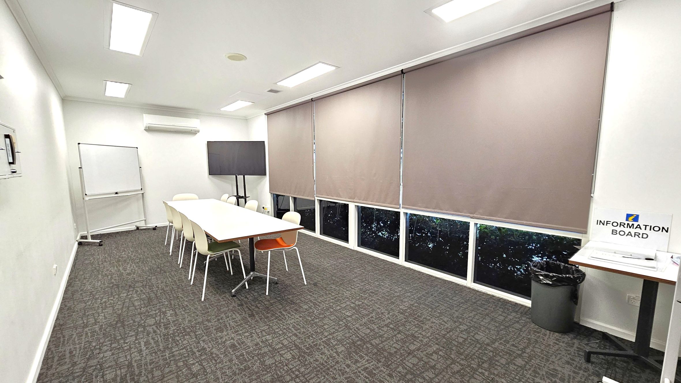 A conference room with a long white table in the centre surrounded by white chairs, one of which is orange. On the left, a whiteboard stands near the wall. The right side features large windows covered with beige blinds. An information board and a bin are positioned in the corner. The floor is carpeted in a grey pattern, and the ceiling has recessed lighting and an air conditioning unit.