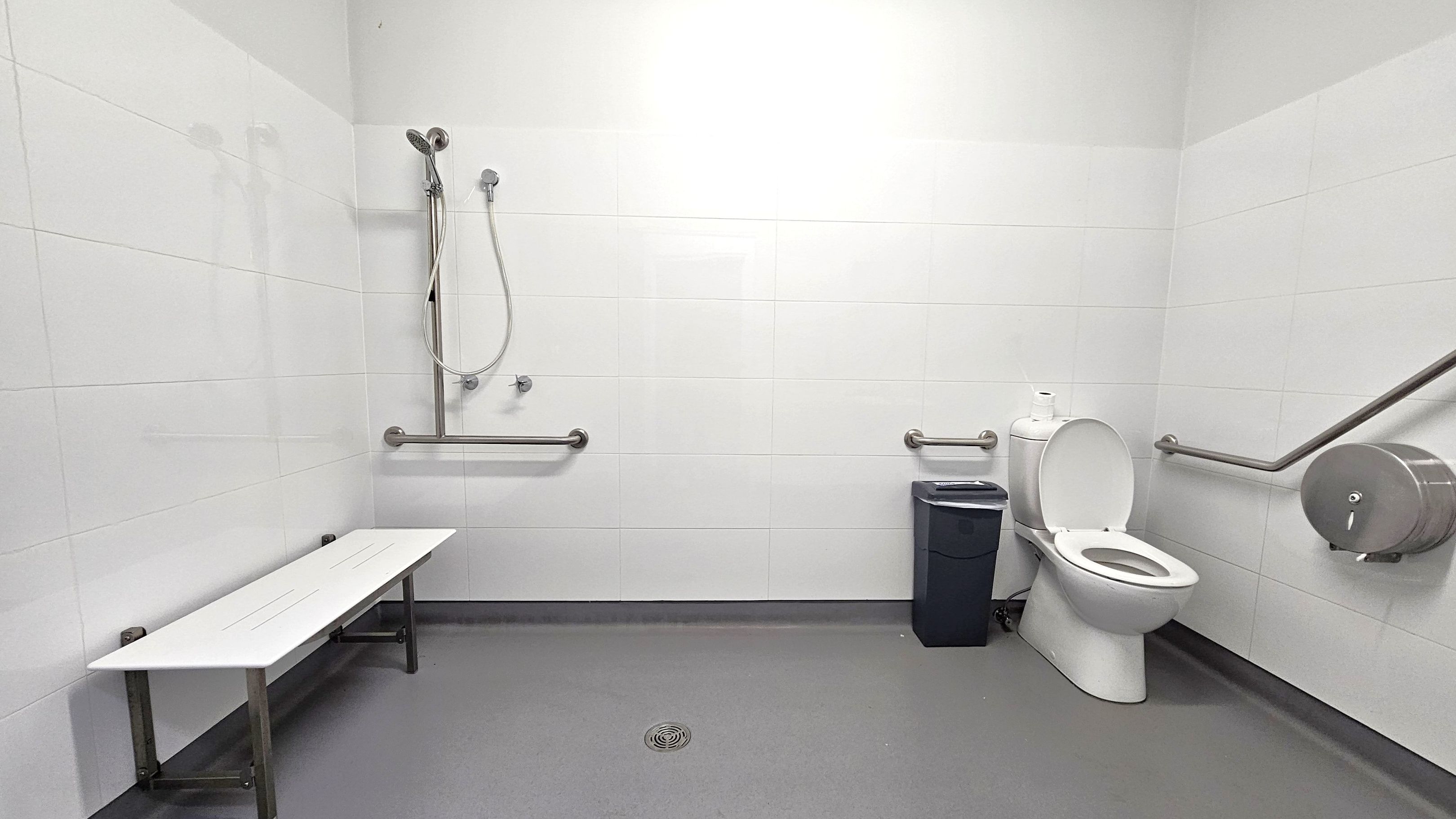 A spacious, accessible bathroom with white tiled walls and a grey floor. On the left, a white bench is mounted to the wall. In the centre, a showerhead with a flexible hose is attached to the wall, accompanied by horizontal grab bars. On the right, a toilet with an open lid is next to a black waste bin. A large toilet paper dispenser and additional grab bars are mounted on the wall near the toilet. A floor drain is visible in the centre.