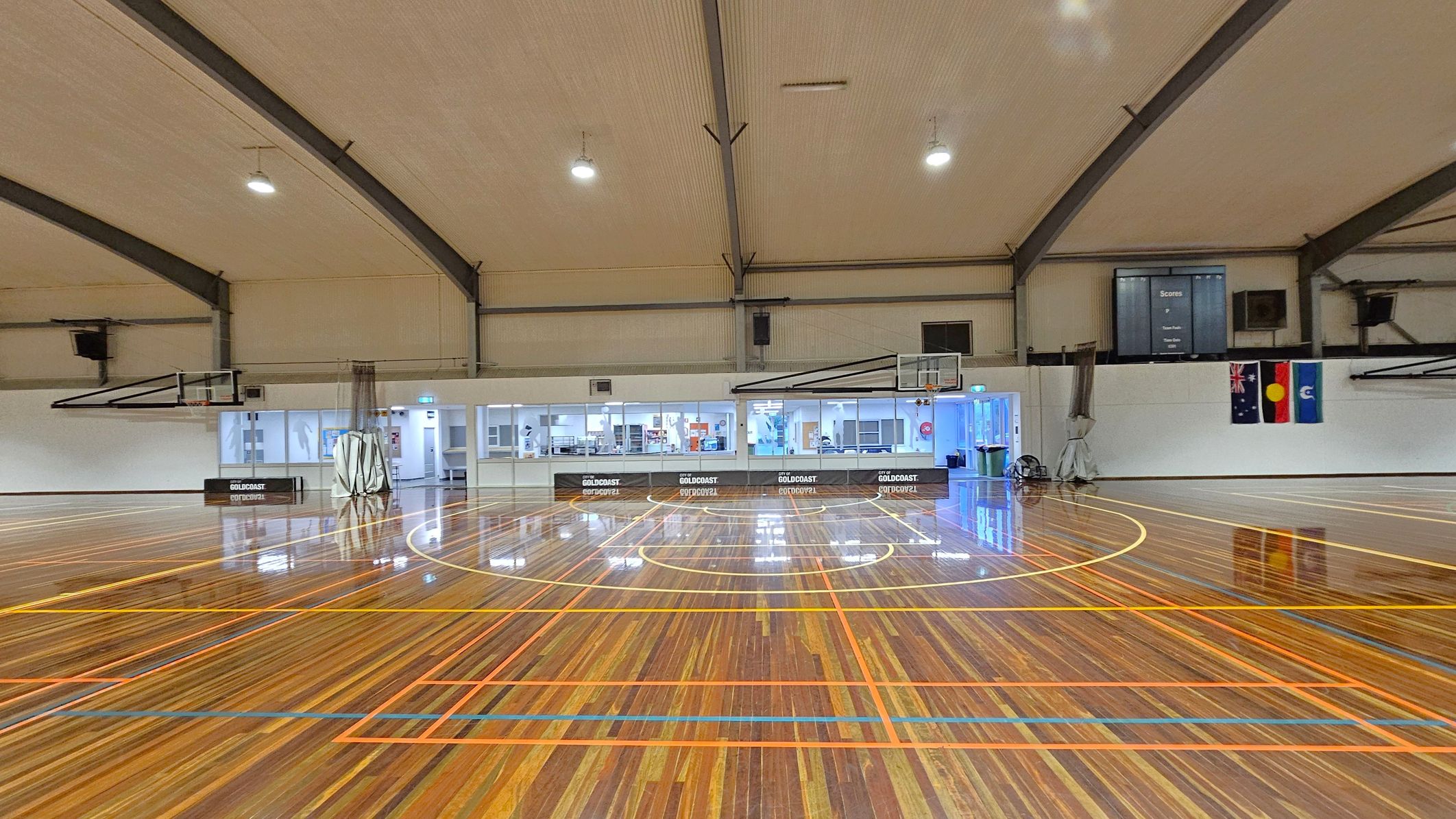 A spacious indoor basketball court with a polished wooden floor featuring orange and yellow lines. Two basketball hoops are visible on either side. The ceiling is high with exposed beams and overhead lights. The back wall has a row of windows and doors, with a scoreboard and three flags displayed. The area is well-lit, and the floor reflects the light, creating a glossy appearance.