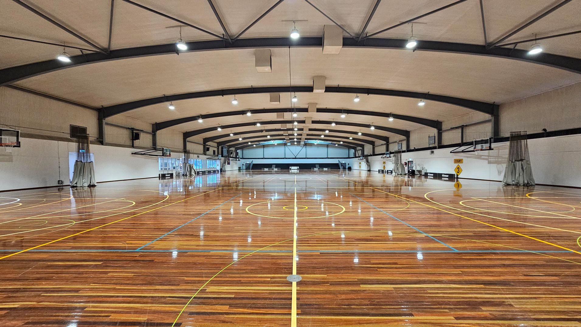 A spacious indoor basketball court with a polished wooden floor marked with yellow and blue lines. The ceiling is high with exposed beams and multiple lights. On both sides, there are basketball hoops and white walls with some windows. A sign on the right indicates "Court 3". The far end of the court has a darker area with additional seating or equipment.