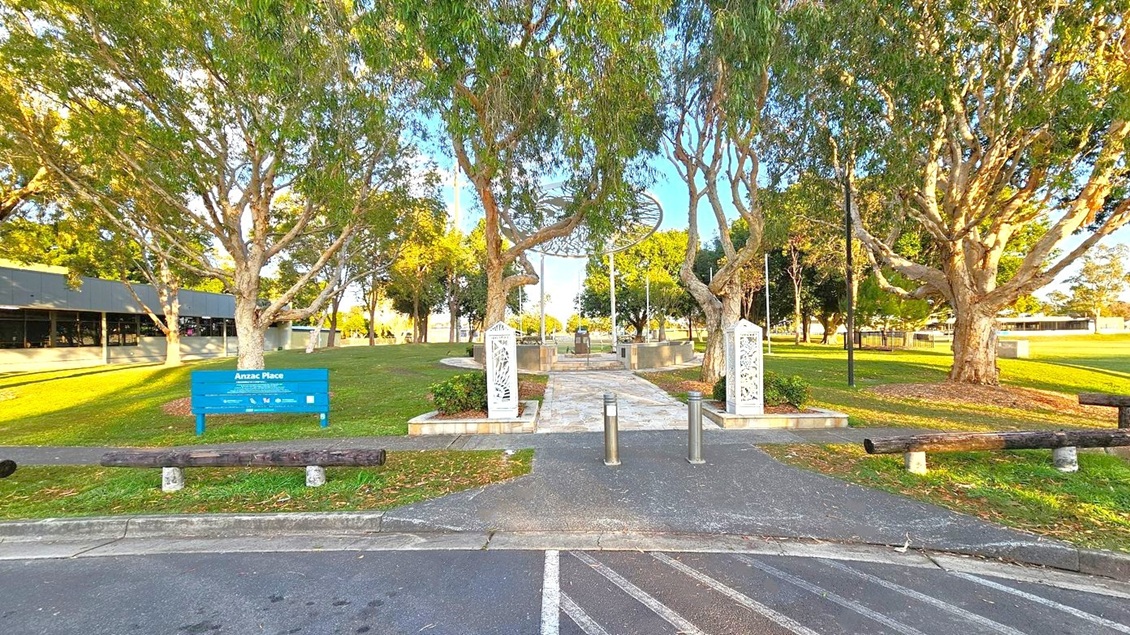 Pathway leading to a memorial area with decorative white pillars on either side. Tall trees with dense foliage line the path, casting shadows on the grass. A blue sign reading "Anzac Place" is on the left, near a modern building with a dark facade. The foreground features a paved area with wooden barriers, and the background shows more trees and open green space.