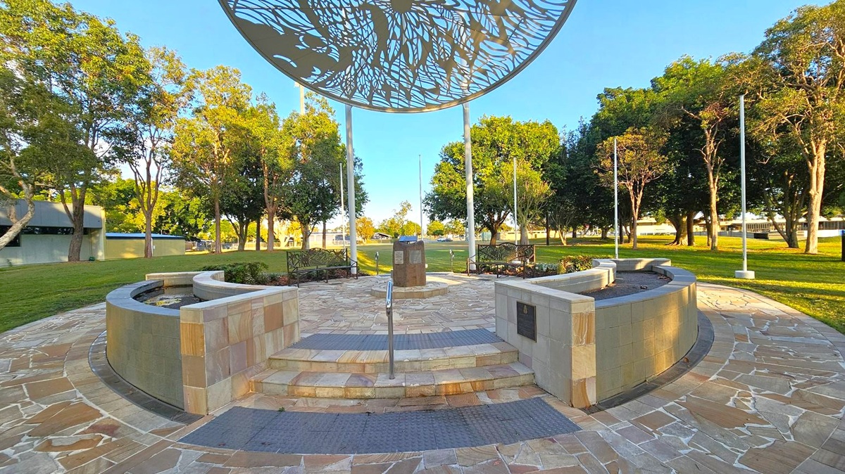 A circular paved area with a central monument surrounded by curved stone benches. Above, a large metal canopy with intricate cut-out patterns is supported by poles. The foreground features stone steps leading up to the monument, with a metal handrail. Benches and flagpoles are positioned around the circle. The background is a grassy park with numerous trees and a few buildings visible in the distance.