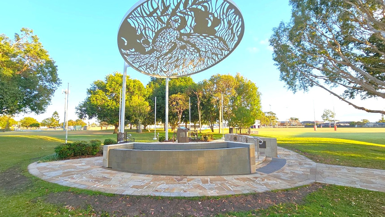 A circular stone platform with a decorative metal canopy featuring intricate patterns is in the centre. The platform is surrounded by a paved walkway. Trees are scattered around the area, with a grassy field extending into the background. Rugby posts are visible in the distance. The sky is clear, and the scene is well-lit, suggesting daytime.