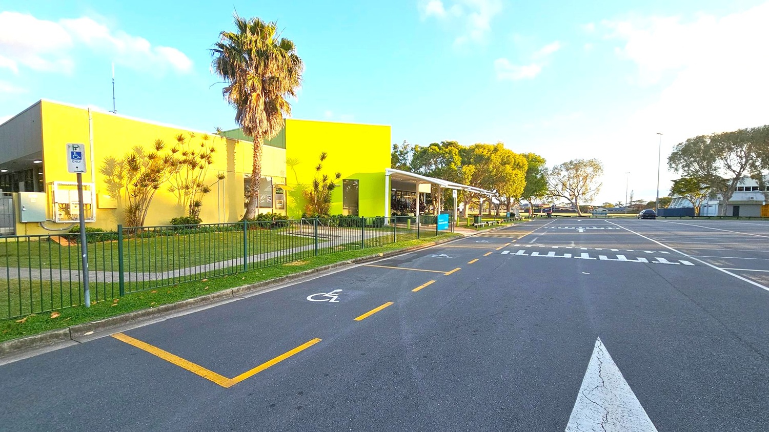 On the left, a yellow building with a bright green section and several windows is surrounded by a grassy area with a palm tree and other plants. A metal fence runs along the grass. A sign indicates a disabled parking space on the road, which is marked with yellow lines. On the right, a row of trees lines the street, and a few cars are parked in the distance. The road features a zebra crossing and directional arrows.