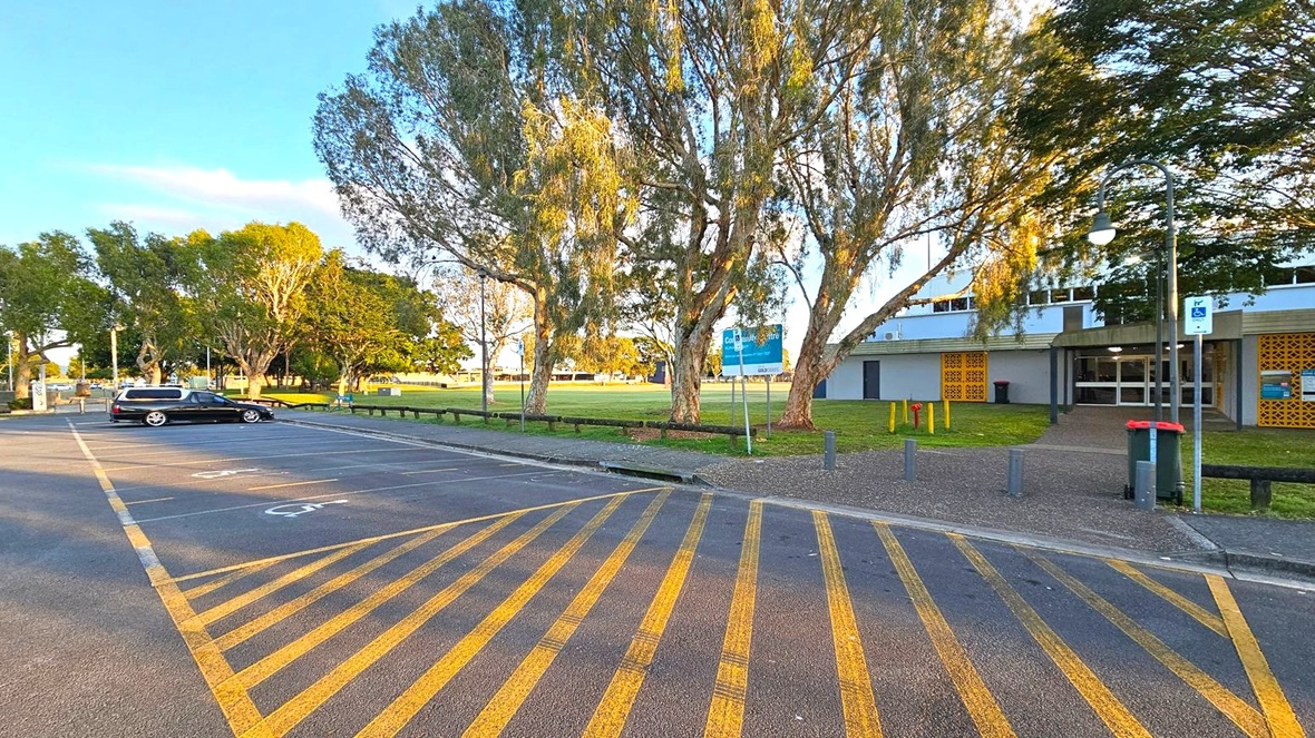 A car park with yellow diagonal lines on the ground is in the foreground. A single black car is parked on the left. In the middle, there are several large trees with green foliage. On the right, a building with a grey facade and yellow decorative panels is visible. A pathway leads to the entrance, and a green bin is nearby. A signpost and a lamp post are also present. The background features more trees and an open grassy area.
