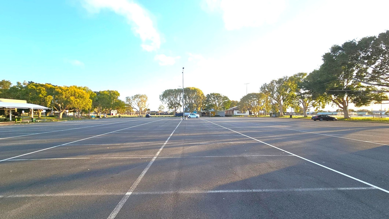 An expansive car park with marked spaces, mostly empty except for a few cars scattered around. Trees line the background, providing a natural border. On the left, a building with a white roof is partially visible. The ground is asphalt, and the area is well-lit, suggesting daylight. A tall light pole stands in the centre, and the scene is open and spacious.