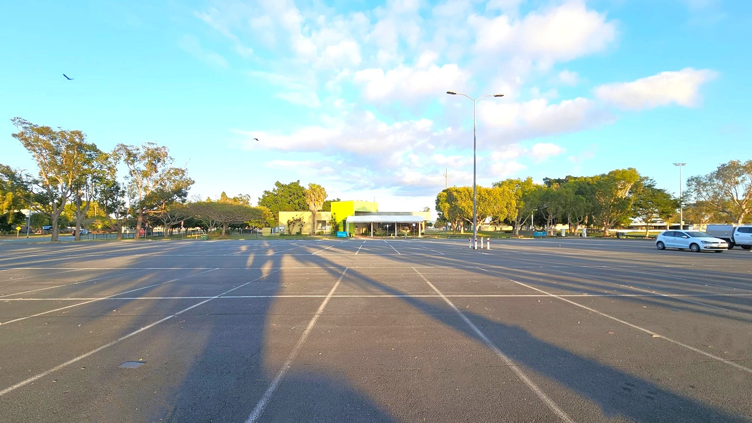 An empty car park with marked spaces and long shadows on the asphalt. In the centre background, a building with a green facade is visible, surrounded by trees. On the right, a few parked cars are seen. The left side features more trees, and birds are flying in the sky. The scene is well-lit, suggesting early morning or late afternoon.