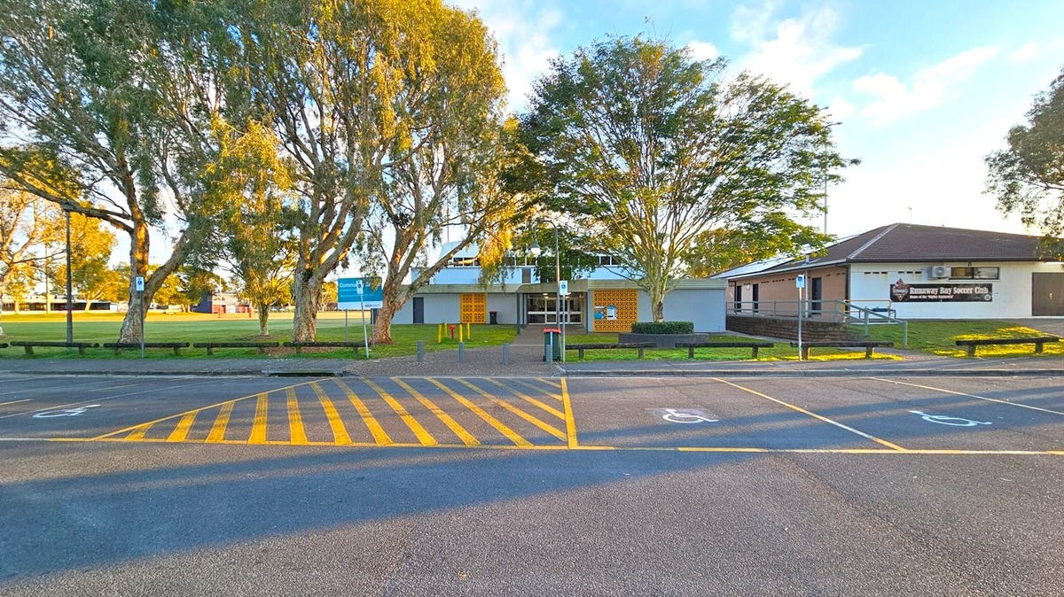 A paved parking area with yellow-striped markings and designated disabled parking spaces in the foreground. In the middle, a building with a sign reading "Runaway Bay Soccer Club" is partially visible on the right. The building is surrounded by trees, with a grassy area and a blue sign on the left. Benches and a pathway lead to the entrance of the building. The scene is well-lit, suggesting daylight.