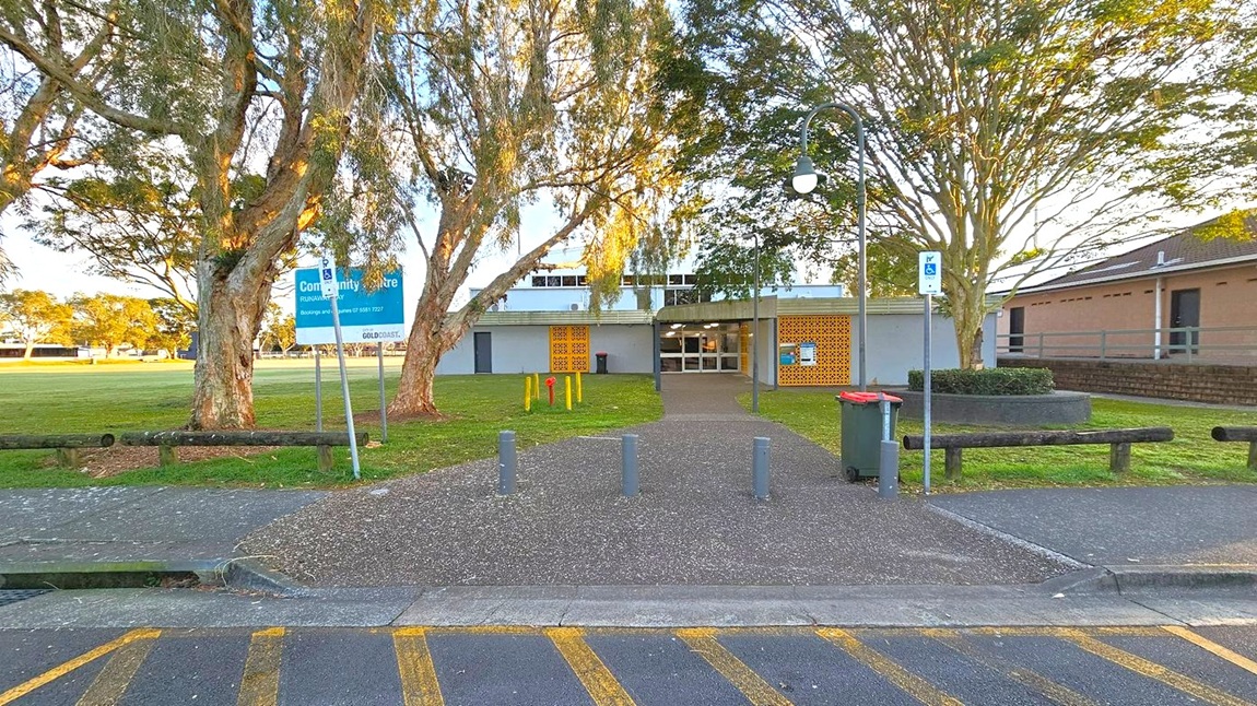 A community centre building is in the centre, with a pathway leading to its entrance. The building has a flat roof and features a mix of glass and decorative brickwork. On the left, there are large trees and a sign indicating the centre's name. On the right, a lamp post and a circular planter with a tree are visible. The foreground has a paved area with bollards and a bin, and the background shows a grassy field and more trees.