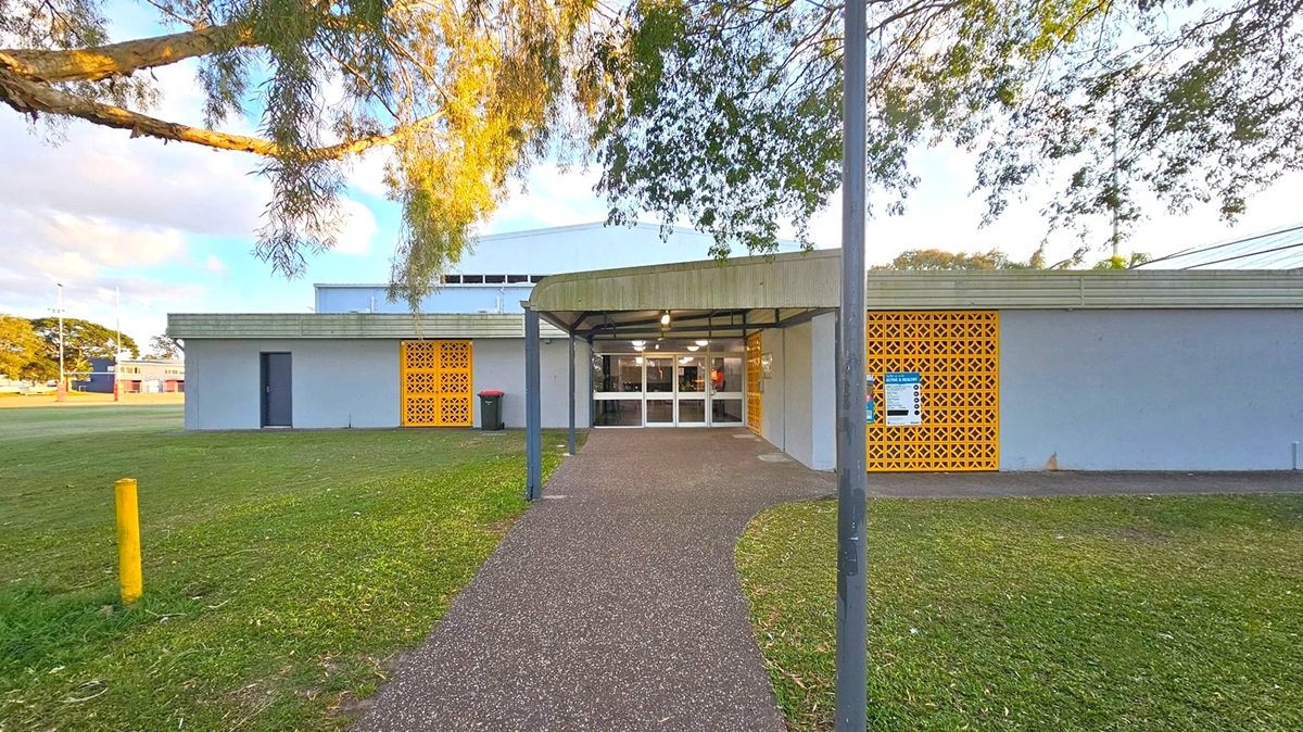 A building with a flat roof and a covered entrance in the centre. The entrance has glass doors and is flanked by yellow lattice panels on both sides. A pathway leads up to the entrance, bordered by grass. A tree branch extends from the left, casting shadows on the building. A yellow bollard is visible on the left side of the grass. The building has a grey exterior with a small dark door on the left.