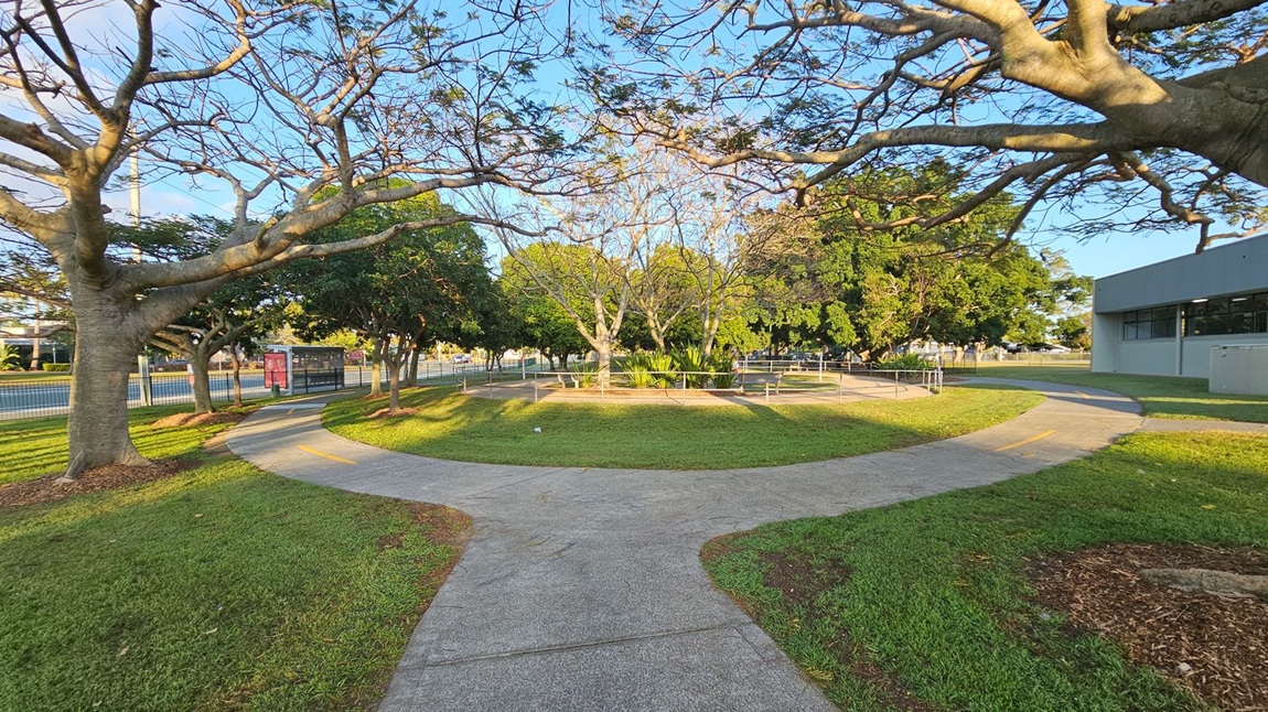A park scene with a concrete path splitting into two directions in the foreground. On the left, large trees with bare branches stretch over the path, casting shadows on the grass. In the background, more trees with dense green foliage are visible. To the right, a grey building with windows is partially visible. A fenced area with plants is in the centre, and a bus stop shelter is seen on the left near the road. The grass is well-maintained, and the sky is clear.