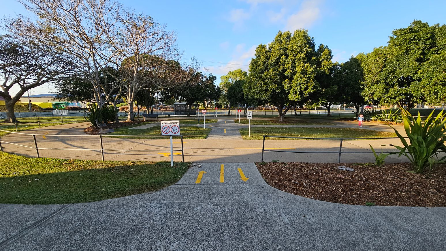 A park with a paved pathway in the centre, marked with yellow arrows and surrounded by grass. Signs along the path include "Wear a Helmet" and traffic symbols. Trees with varying foliage are scattered throughout, with some bare branches on the left and lush green on the right. A child is visible in the distance on the right. The foreground has a concrete path leading into the park, bordered by a low metal fence and mulch with plants.