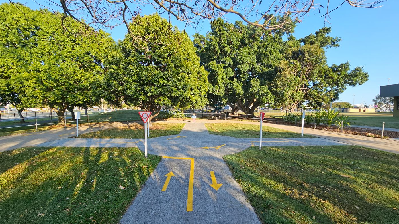 A paved path with yellow directional arrows leads to a small roundabout with give way signs. The path is surrounded by green grass and large, leafy trees. In the background, there is a fence and more trees. On the right, there are some small plants and a building partially visible. The sky is clear and blue, and the shadows of the trees stretch across the grass and path.
