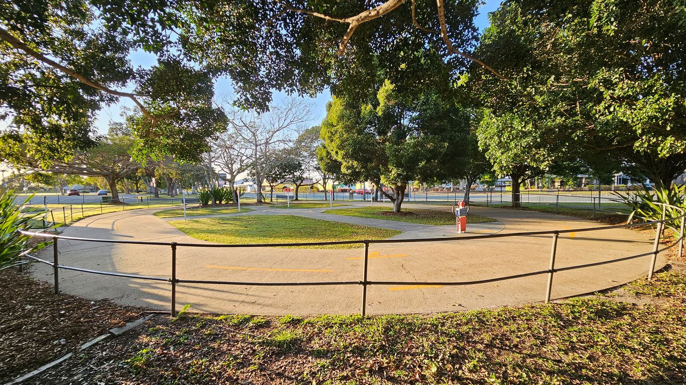 A park with a circular paved path surrounded by a black metal railing. The path is bordered by grass and trees, with some trees having dense green foliage and others bare. A small red and white post is visible near the centre. In the background, there is a fence and a road with parked cars. The ground is covered with fallen leaves, and the sunlight casts shadows from the trees.