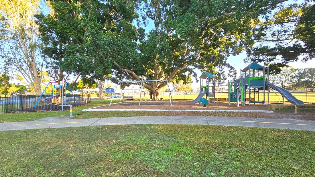 A playground with a large tree in the centre, providing shade. On the left, there's a fenced area with a swing set. In the middle, a metal swing frame stands on a mulch-covered ground. On the right, a play structure with slides and climbing features is visible. The ground is grassy with a concrete path running across the foreground. The background shows more trees and a fence enclosing the area.