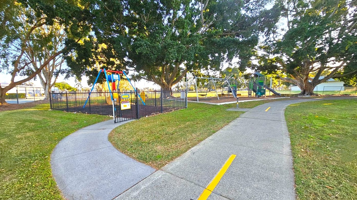A park with a playground featuring a fenced area on the left containing a swing set. A paved path with a yellow line runs through the centre, dividing the grassy area. On the right, there is another swing set and a climbing structure with slides. Large trees with dense foliage provide shade throughout the area. The background includes more trees and a glimpse of a building. The ground is a mix of grass and mulch under the play equipment.