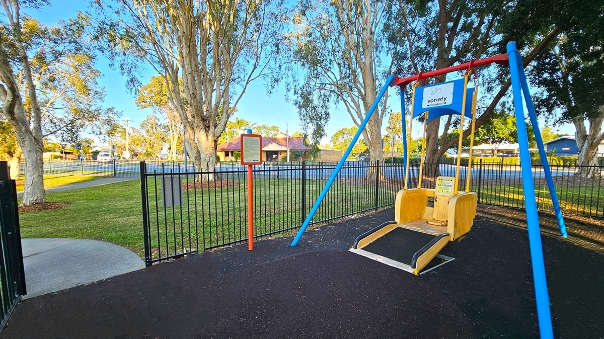 A playground with a swing designed for accessibility is on the right, featuring a blue and red frame. In the centre, a signpost with information stands on a grassy area. The background has a black metal fence and several tall trees with light-coloured trunks. Beyond the fence, a building with a red-tiled roof is visible. The ground is covered with dark rubber matting, and a concrete path leads to the gate on the left.