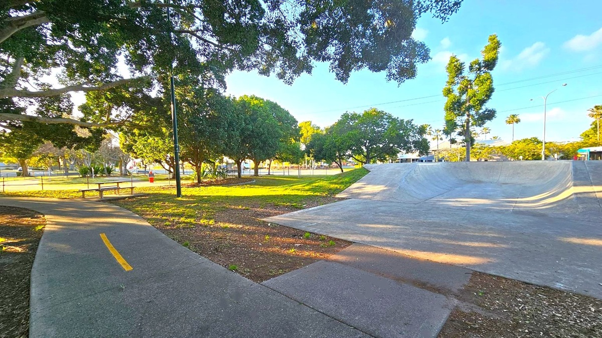 A skate park with a concrete ramp on the right. On the left, a curved path with a yellow line runs through a grassy area. Several large trees provide shade, and a bench is visible near the path. In the background, more trees and a fence can be seen, along with a few streetlights and a building. The ground is a mix of grass and dirt, and the area is well-lit by natural light.