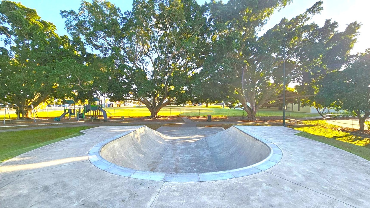 Concrete skate bowl in the foreground with a smooth surface. In the background, large trees with dense foliage provide shade. To the left, a playground with slides and swings is visible. The ground is grassy, and a fence runs along the back. A pathway curves around the skate area, and a lamppost stands to the right. The scene is well-lit, suggesting daylight.