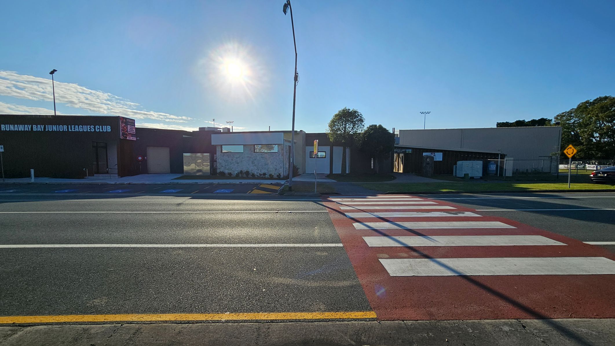 A street scene with a zebra crossing in the foreground, leading to a road with multiple lanes. On the left, a building with the sign "Runaway Bay Junior Leagues Club" is visible. In the centre, a small building with a stone facade and a tree nearby. On the right, a larger building with a plain exterior. A streetlamp stands in the middle, and a road sign is visible on the right. The sun is low in the sky, casting long shadows.