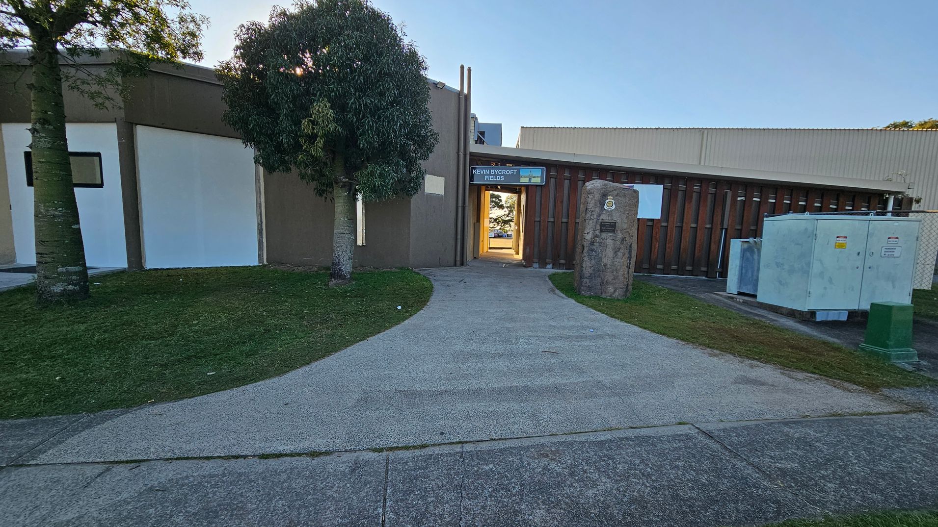 A pathway leads to a building entrance with a sign above it. On the left, there are two trees and a grassy area. On the right, a large stone with a plaque stands near utility boxes. The building has a brown and white exterior with vertical wooden slats. The ground is paved with concrete, and the area is well-maintained.