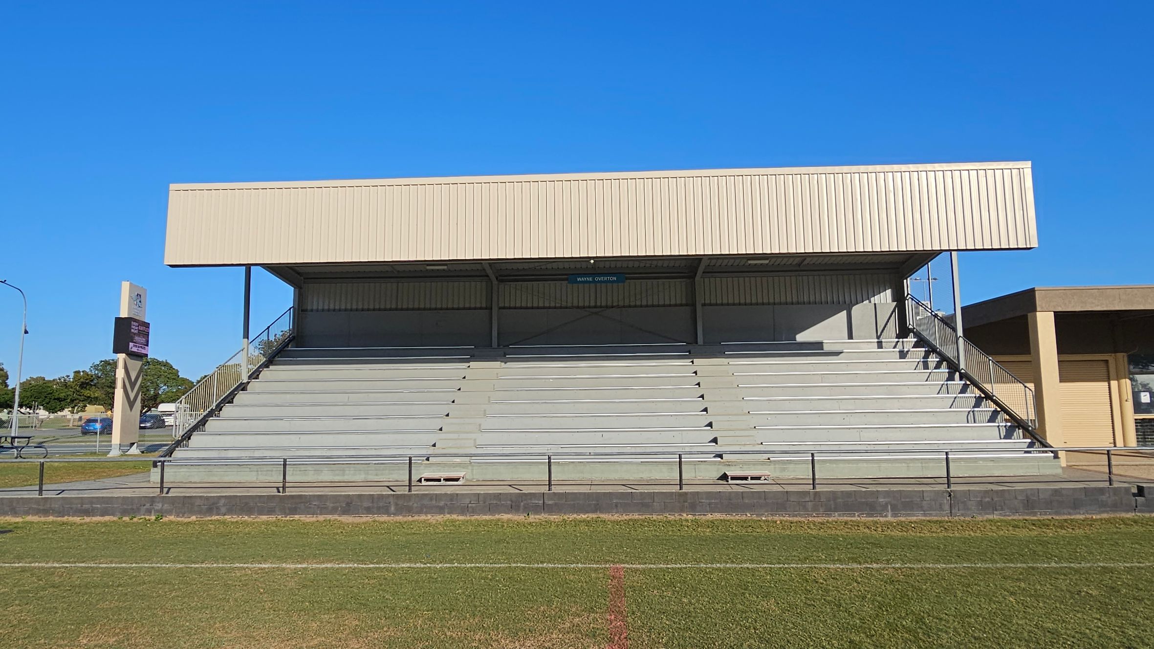 A set of outdoor bleachers with a metal roof and grey seating. The structure is elevated with stairs on both sides. In the foreground, there is a grassy field with a red line marking. To the left, a tall signpost is visible near a road with parked cars and trees. On the right, part of a building with a roller door is visible. The sky is clear and blue.