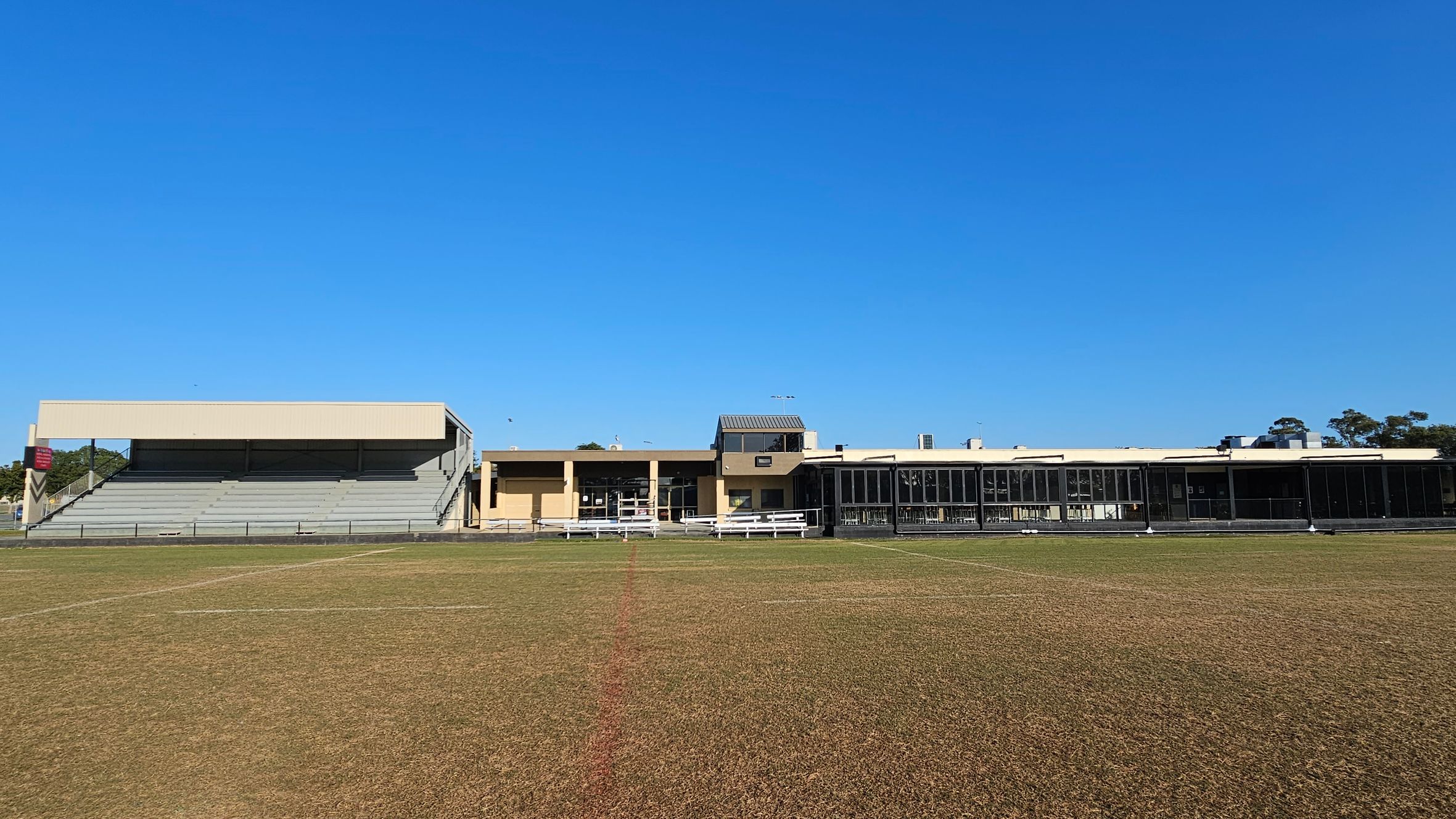 A sports field with a patchy grass surface in the foreground. In the background, there is a large building complex. On the left, there is a covered grandstand with tiered seating. In the centre, a beige building with a small entrance and windows. On the right, a long structure with large glass windows. The sky is clear and blue.