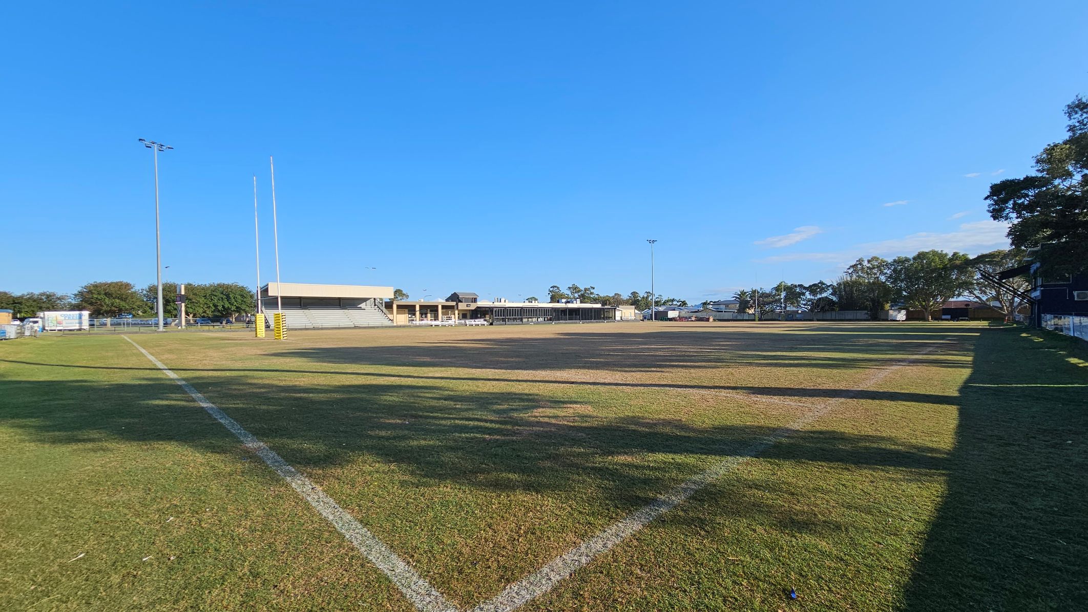 A rugby field with goalposts on the left, surrounded by a grassy area. The field has a worn, patchy surface. In the background, there are stands and buildings, with trees scattered around. Tall floodlights are visible on both sides. The right side has a shadowed area with more trees and a fence. The sky is clear and blue.
